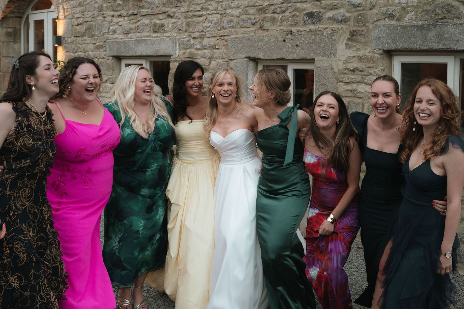 A group of wedding guests in colorful dresses gather for a celebratory photo outside a stone building.