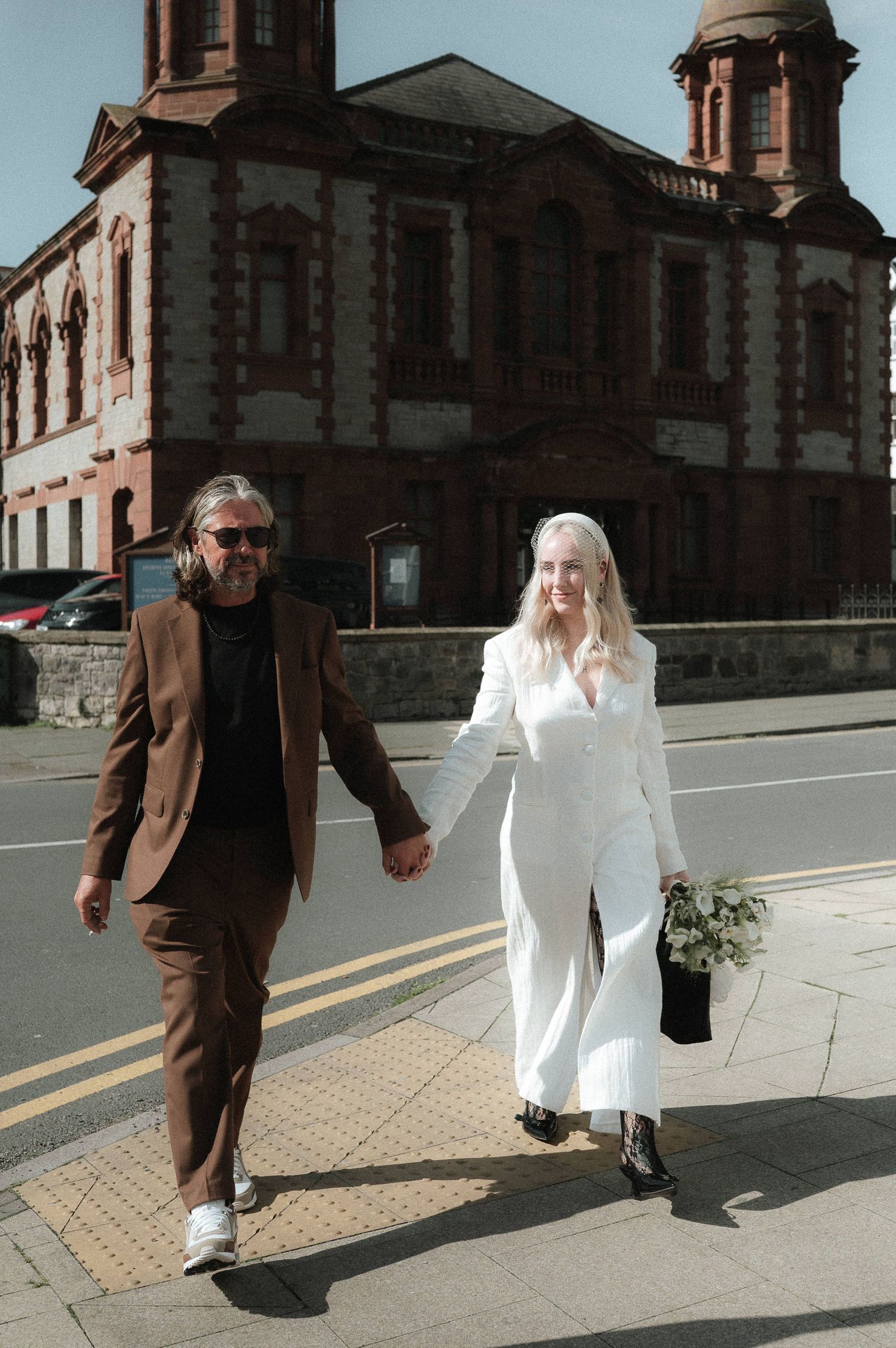A couple in contrasting brown and white outfits walking together on a sidewalk near a brick building.