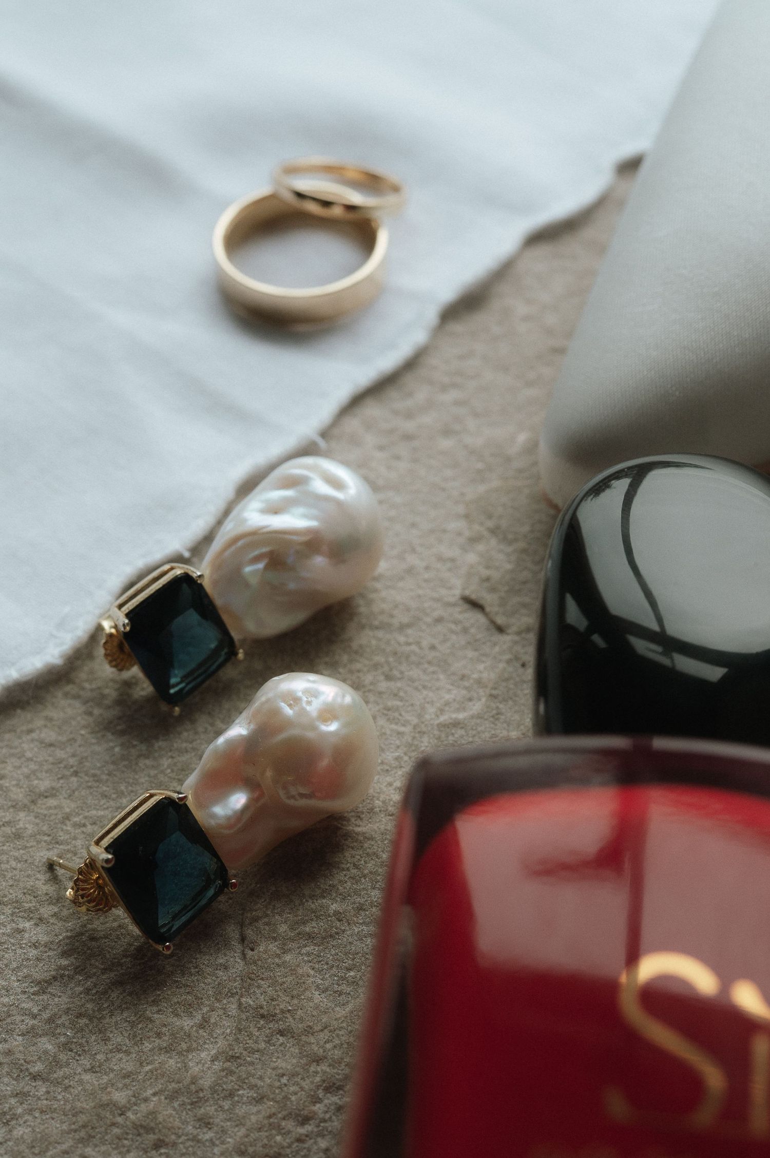Pearl earrings with emerald stones and gold wedding bands displayed on a neutral surface next to a red perfume bottle.