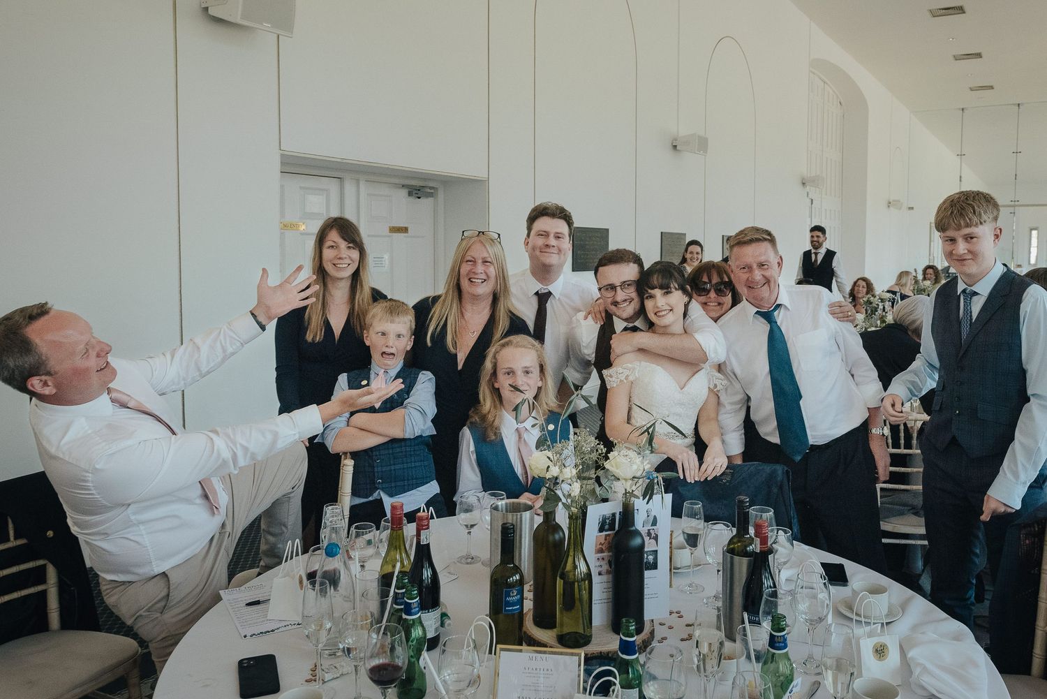 Wedding reception guests gathered around a long table with wine bottles and glassware.