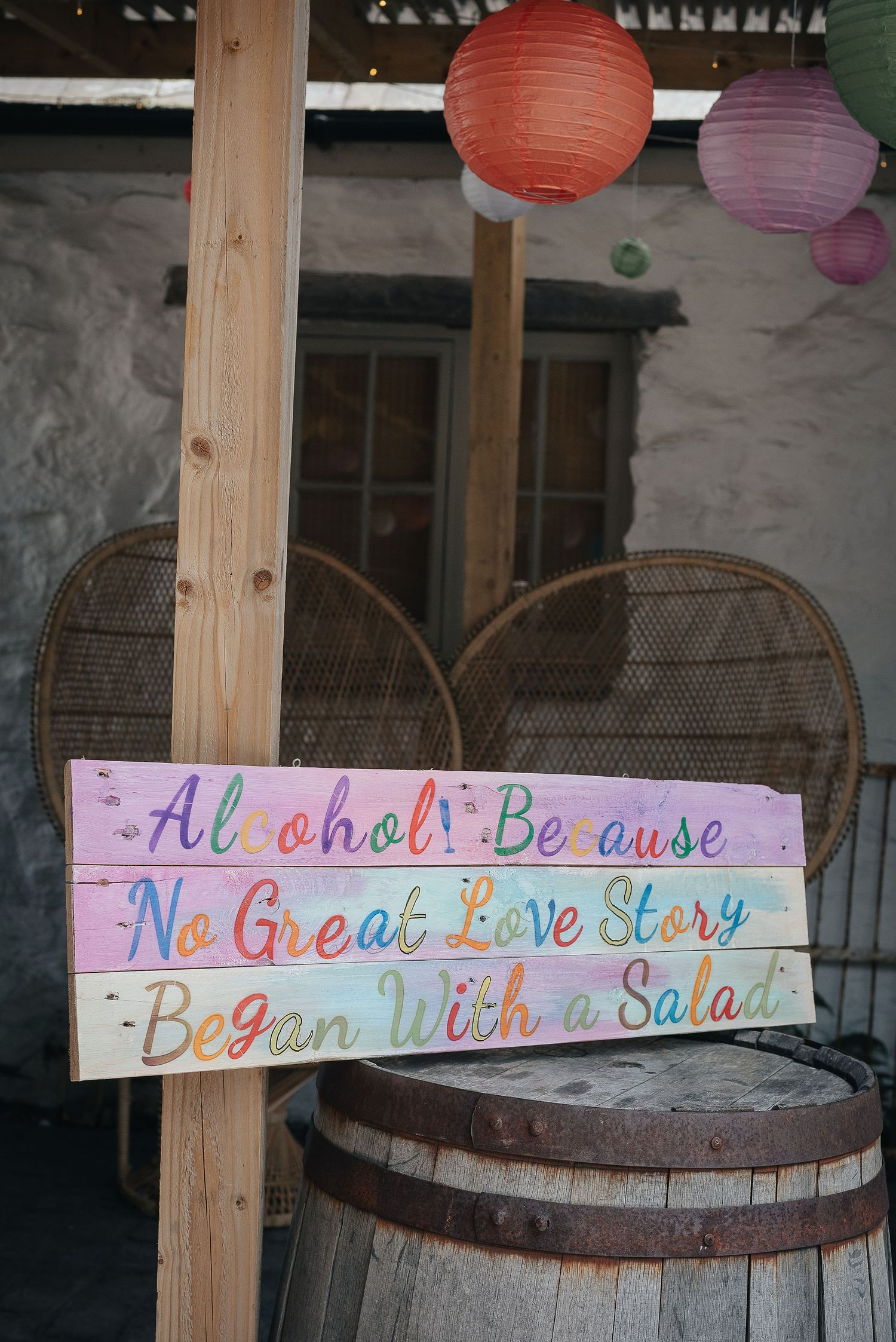 Colorful wedding sign with paper lanterns reading 'Alcohol Because No Great Love Story Began With a Salad'.
