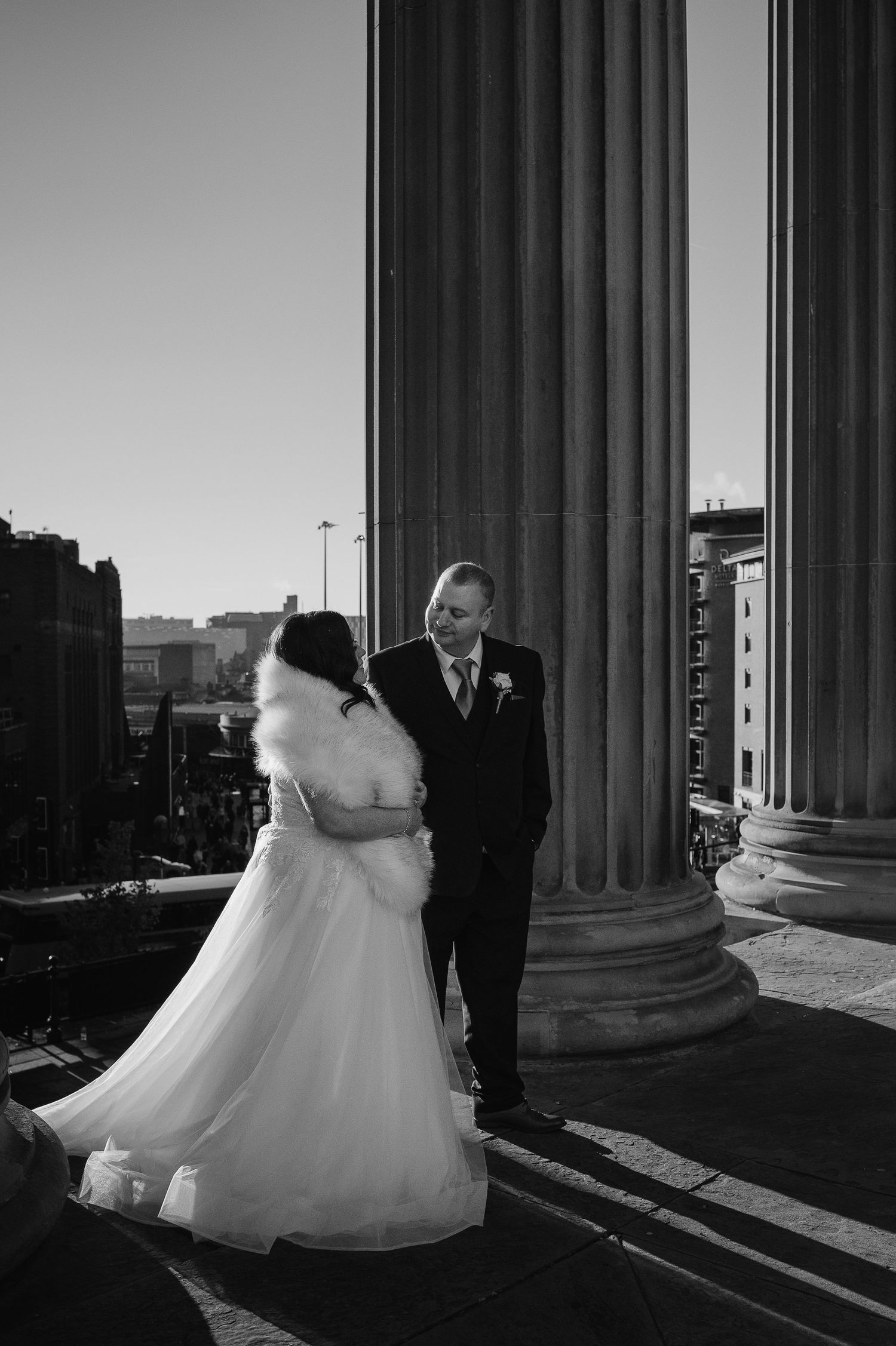 A dramatic black and white wedding portrait framed by classical columns against a city backdrop.
