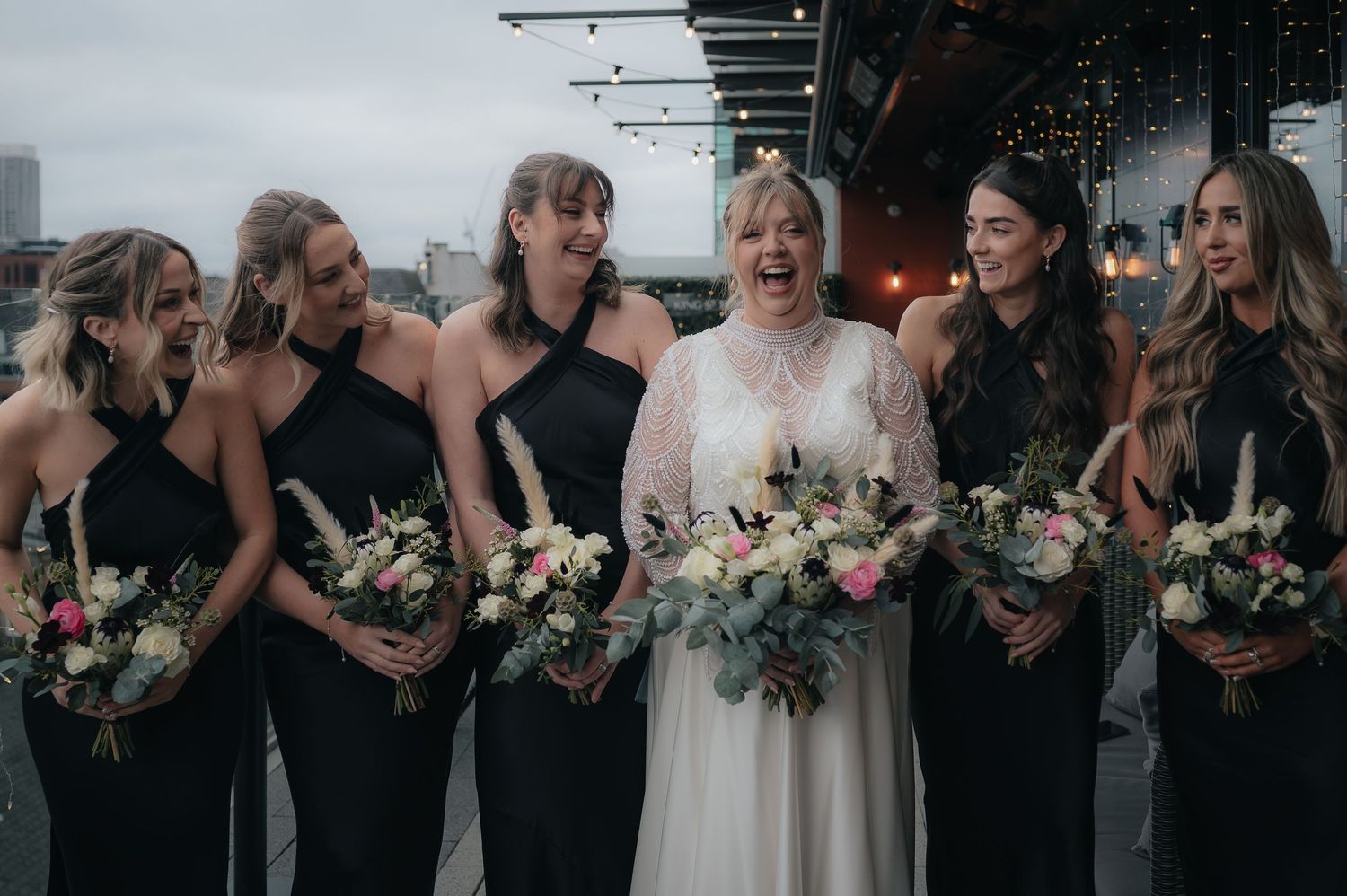 Wedding party in black dresses poses with fresh floral bouquets next to person in white dress during outdoor celebration.