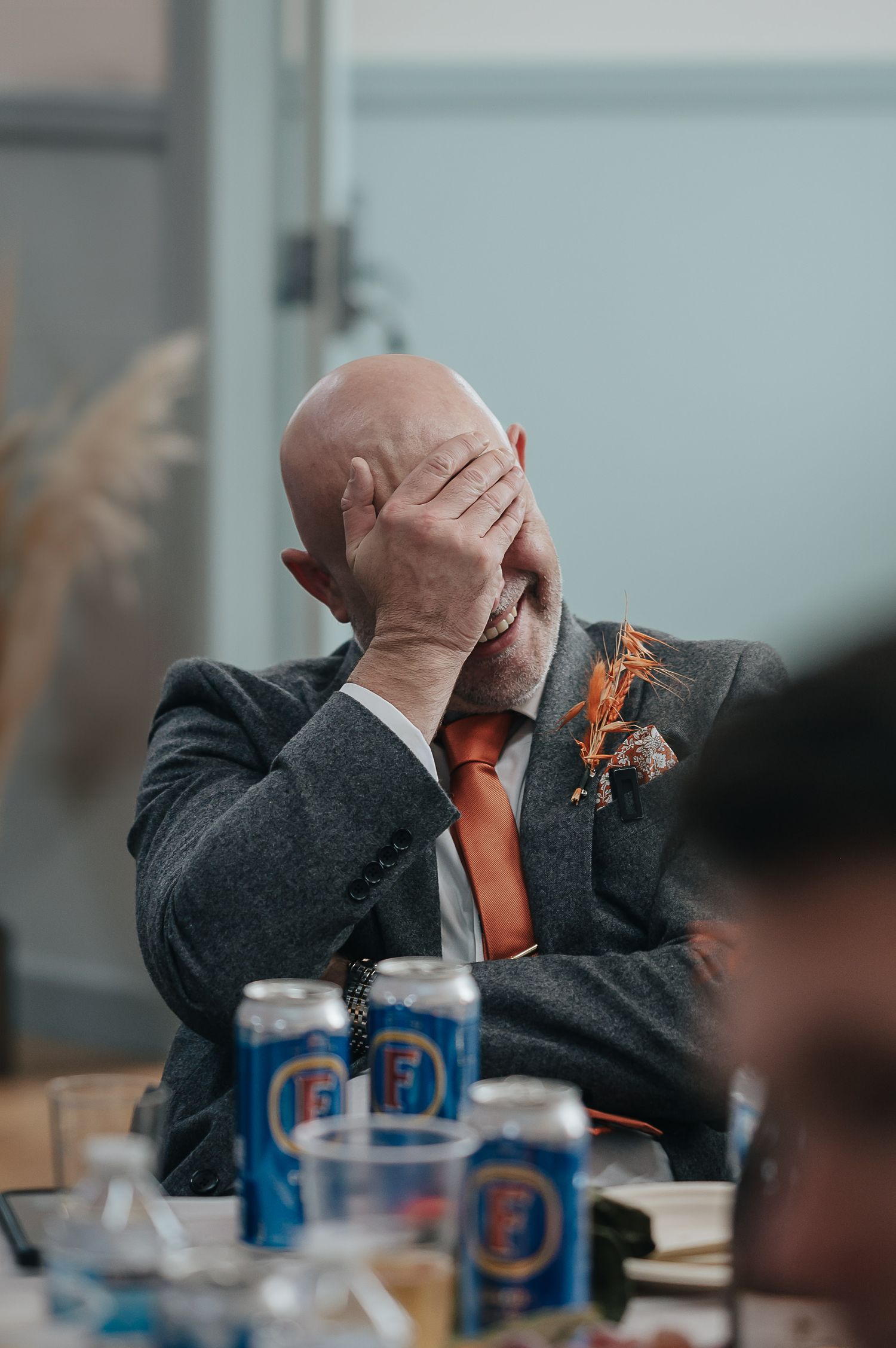 Wedding guest in grey suit with orange tie covers face in an emotional moment during reception.