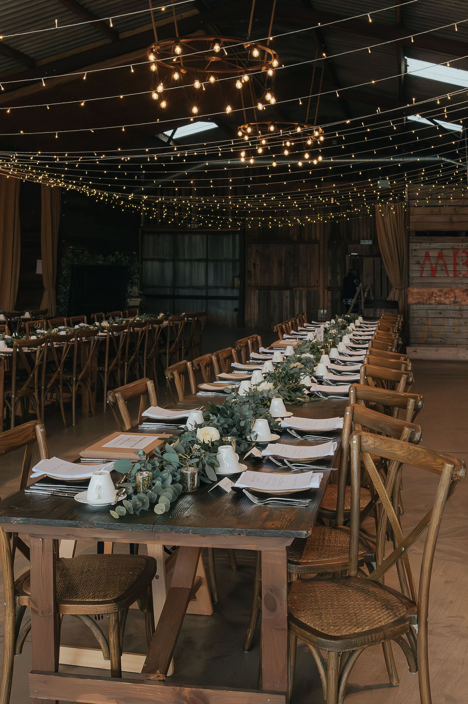Long wooden farm tables set for wedding reception under twinkle lights in rustic venue.