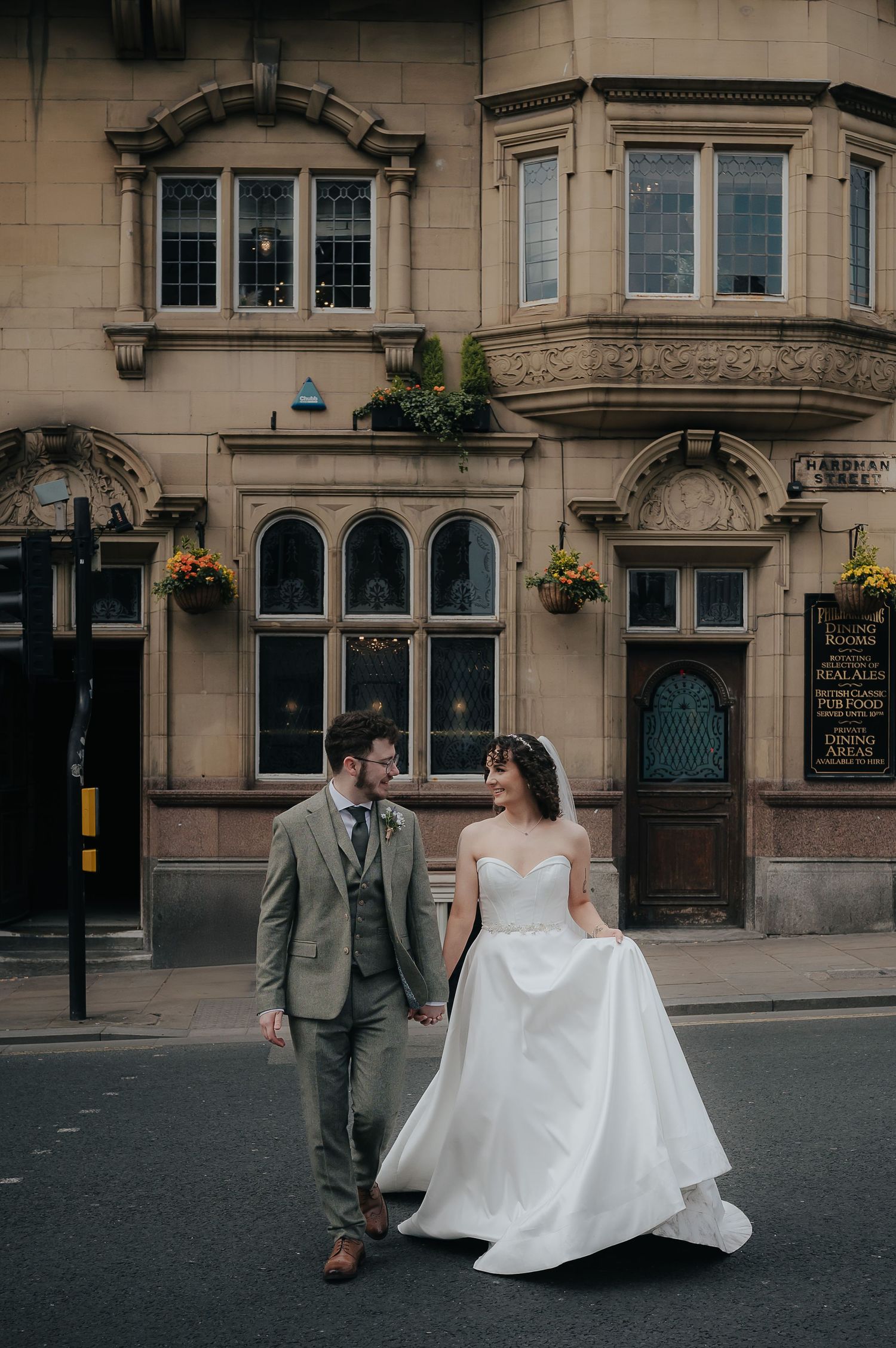 A couple walks together on a city street with ornate historic architecture and hanging flower baskets in the background.