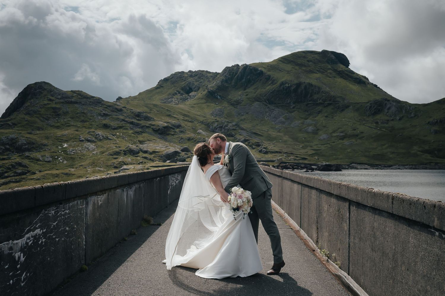 Romantic wedding portrait on mountain dam with dramatic cloudy backdrop.