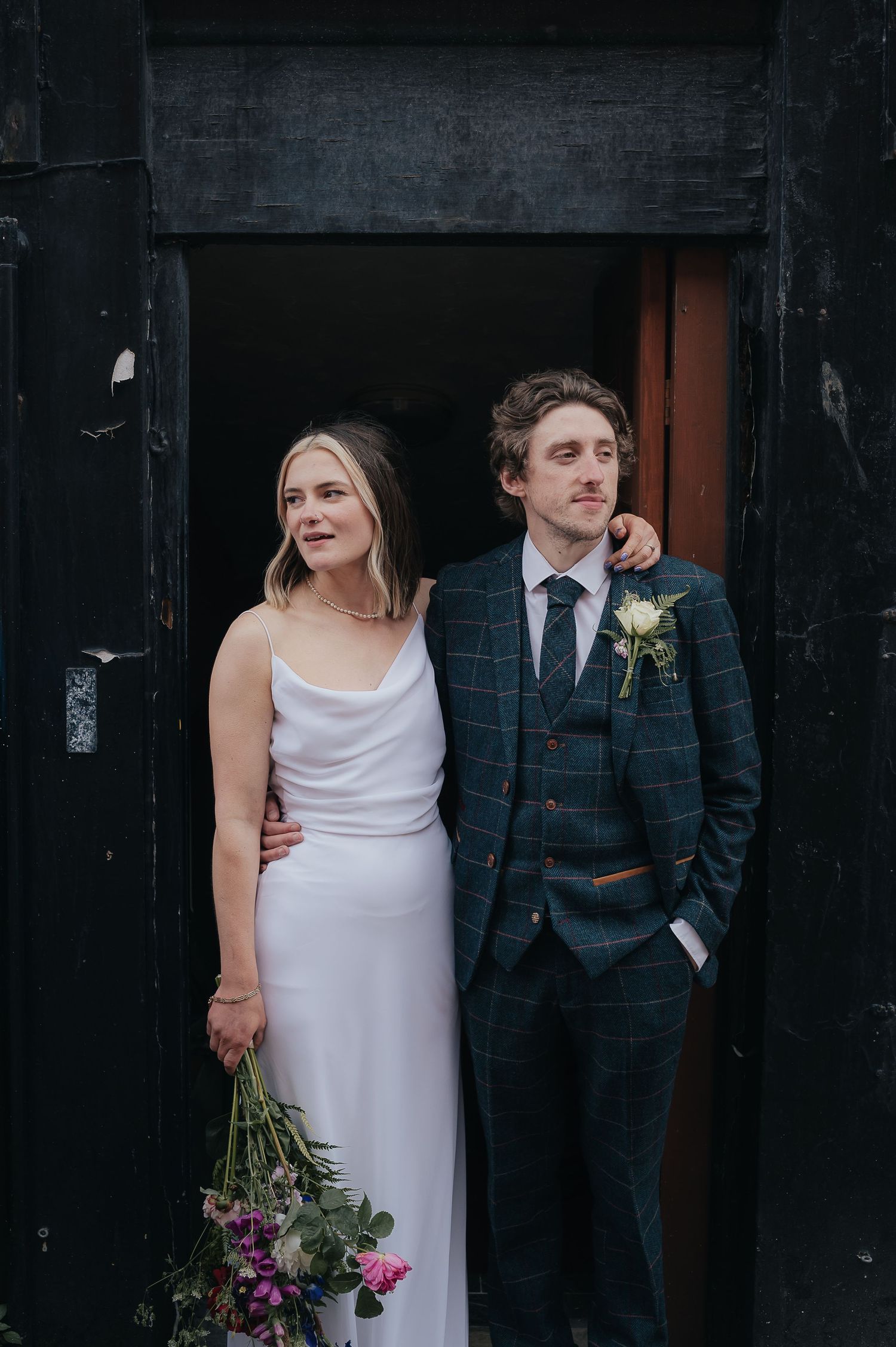 Newlyweds pose in front of a dark doorway, bride in white dress and groom in plaid suit.