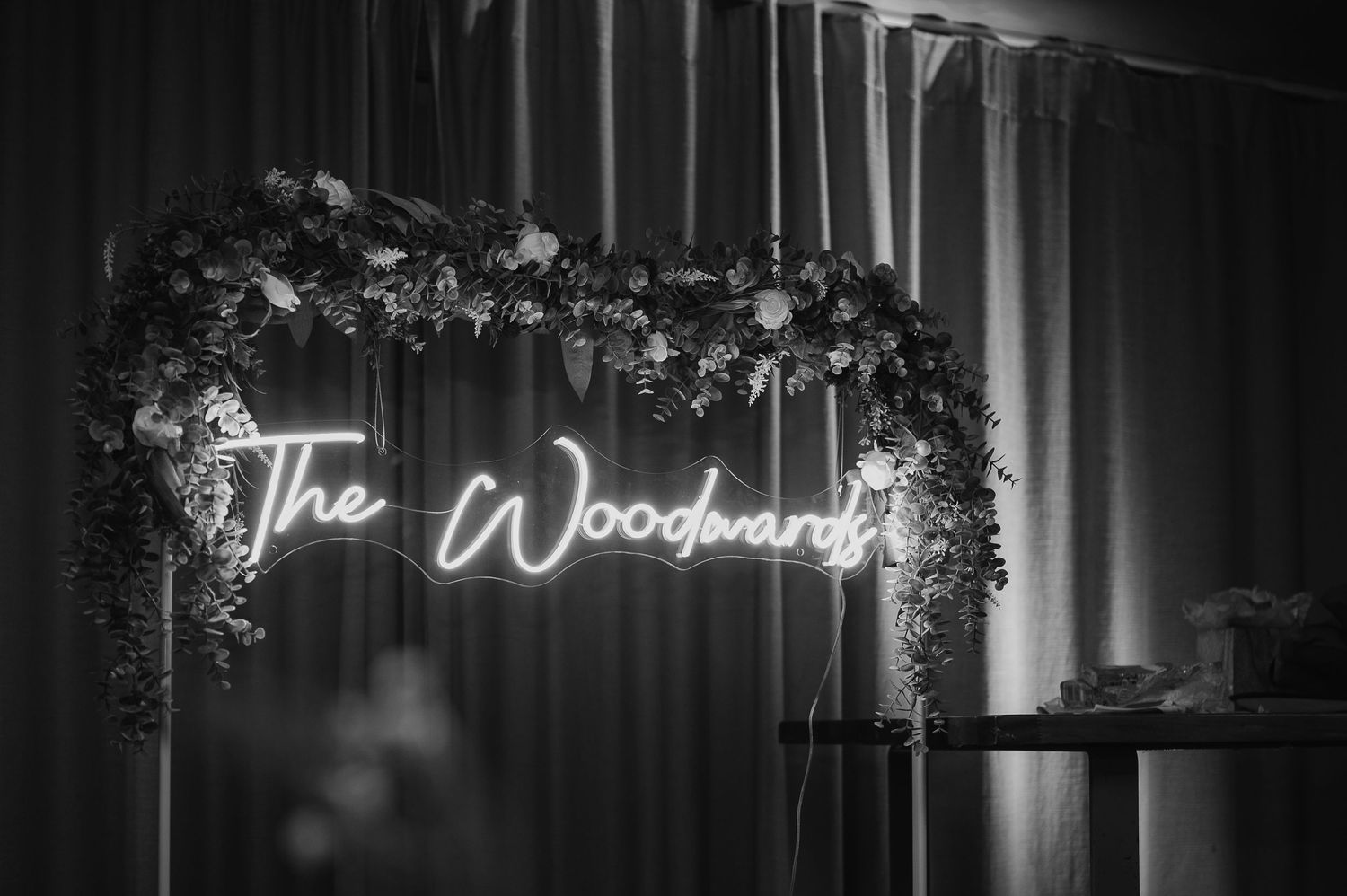 A moody black and white shot of 'The Woodwards' neon sign surrounded by flowers.