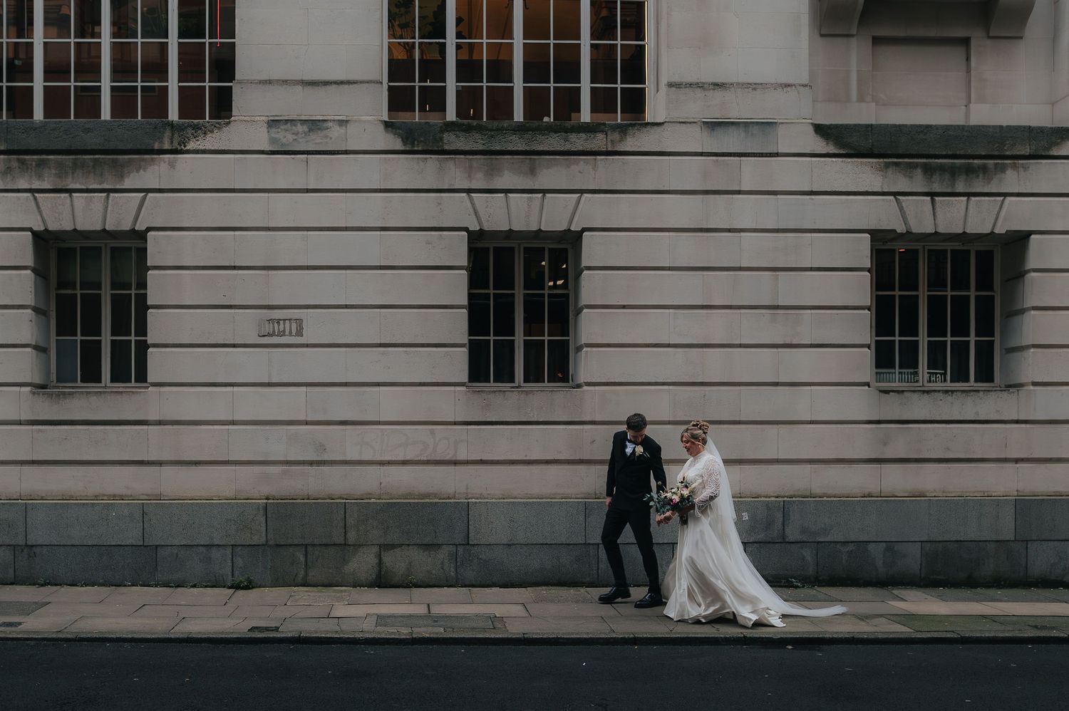 A couple in wedding attire stands against a stone building with classical architecture and barred windows.