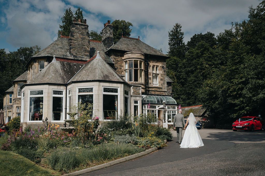 Bride in white dress walking past historic stone mansion with bay windows.
