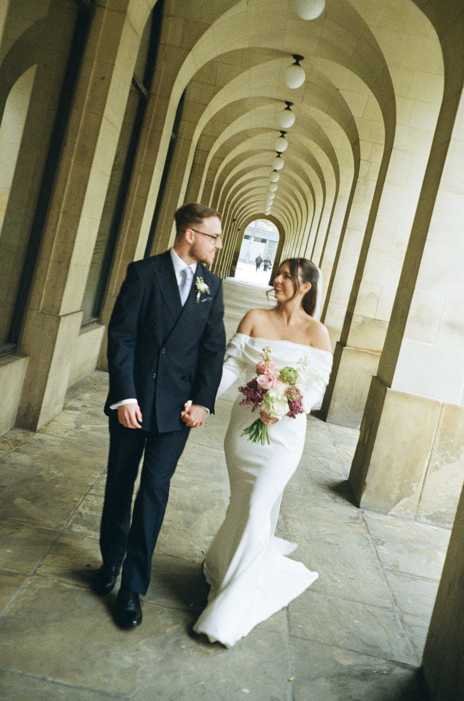 A bride and groom walk hand-in-hand through an elegant arched colonnade, she holds a pink bouquet in a white off-shoulder gown.