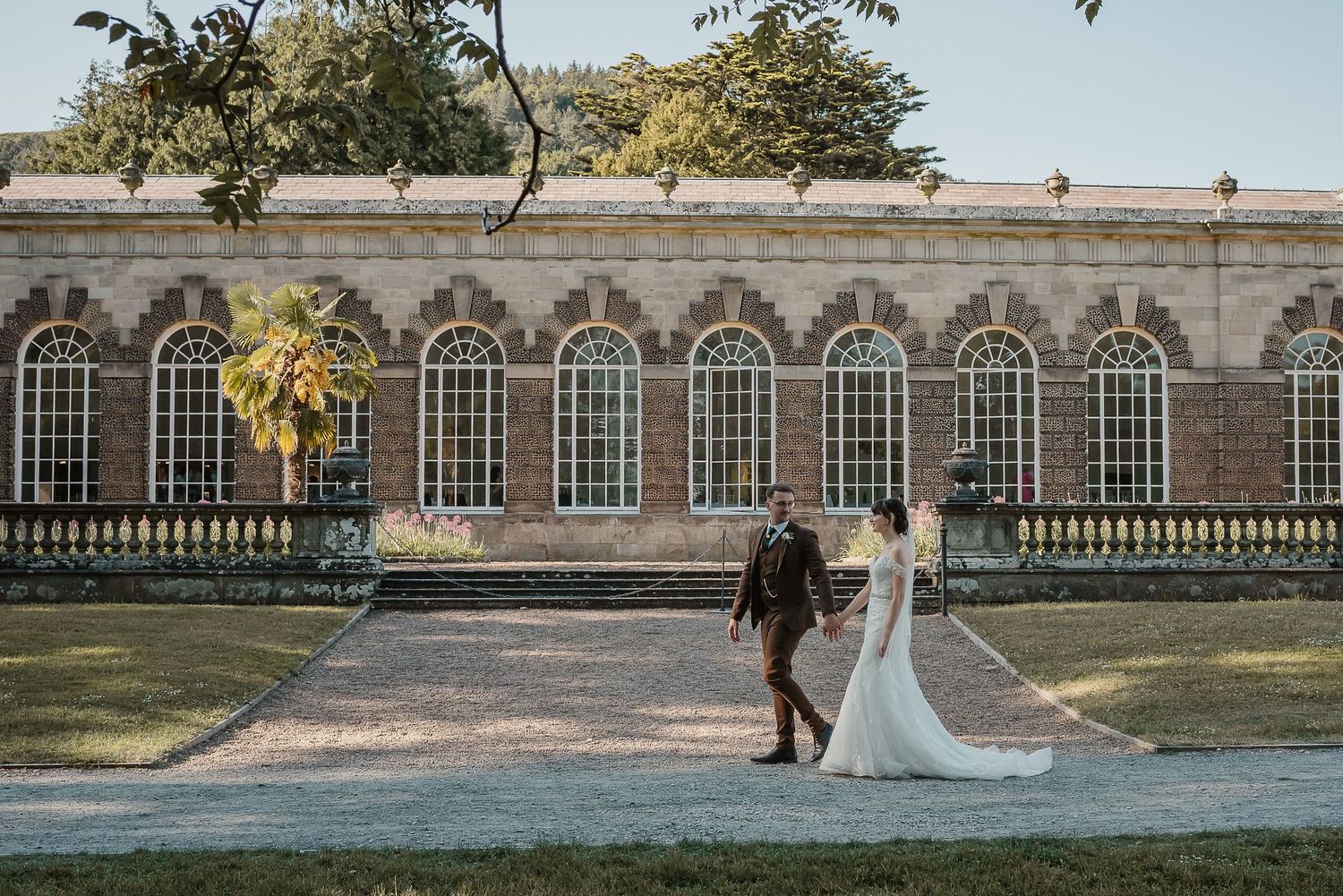 Elegant couple walking near historic building with arched windows and landscaped grounds.