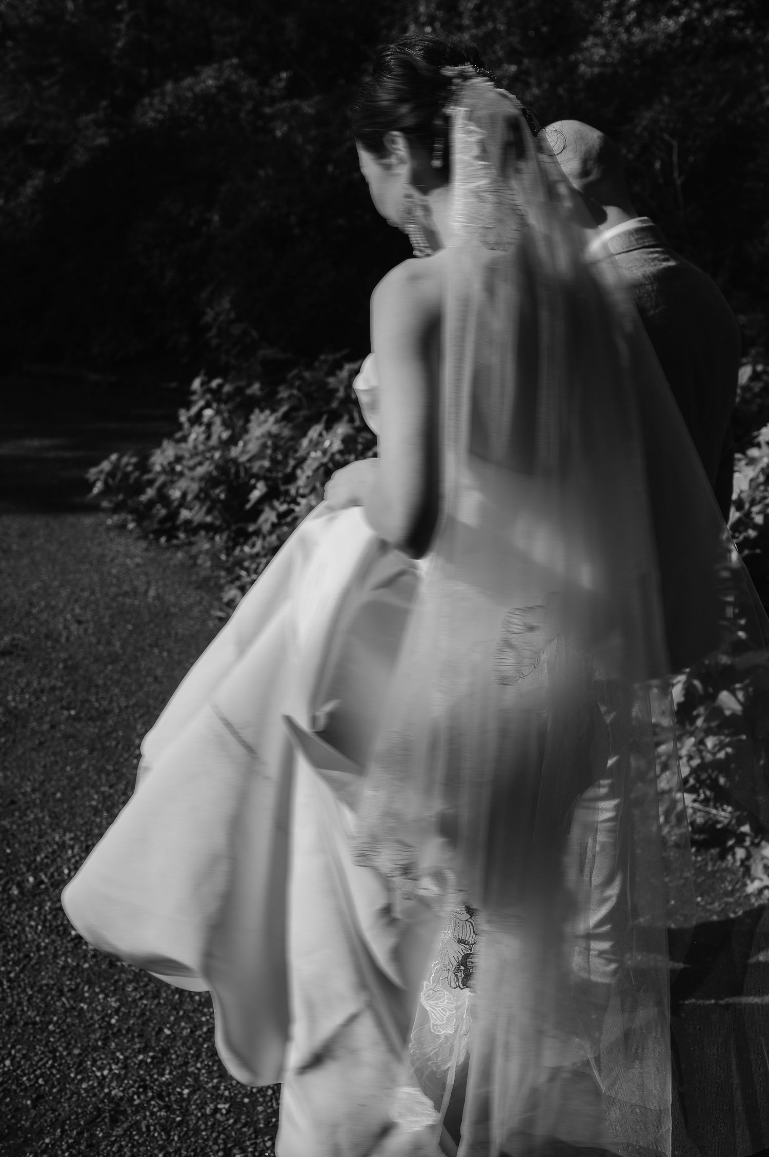 Artistic black and white shot of a bride's dress and flowing veil in a garden setting.