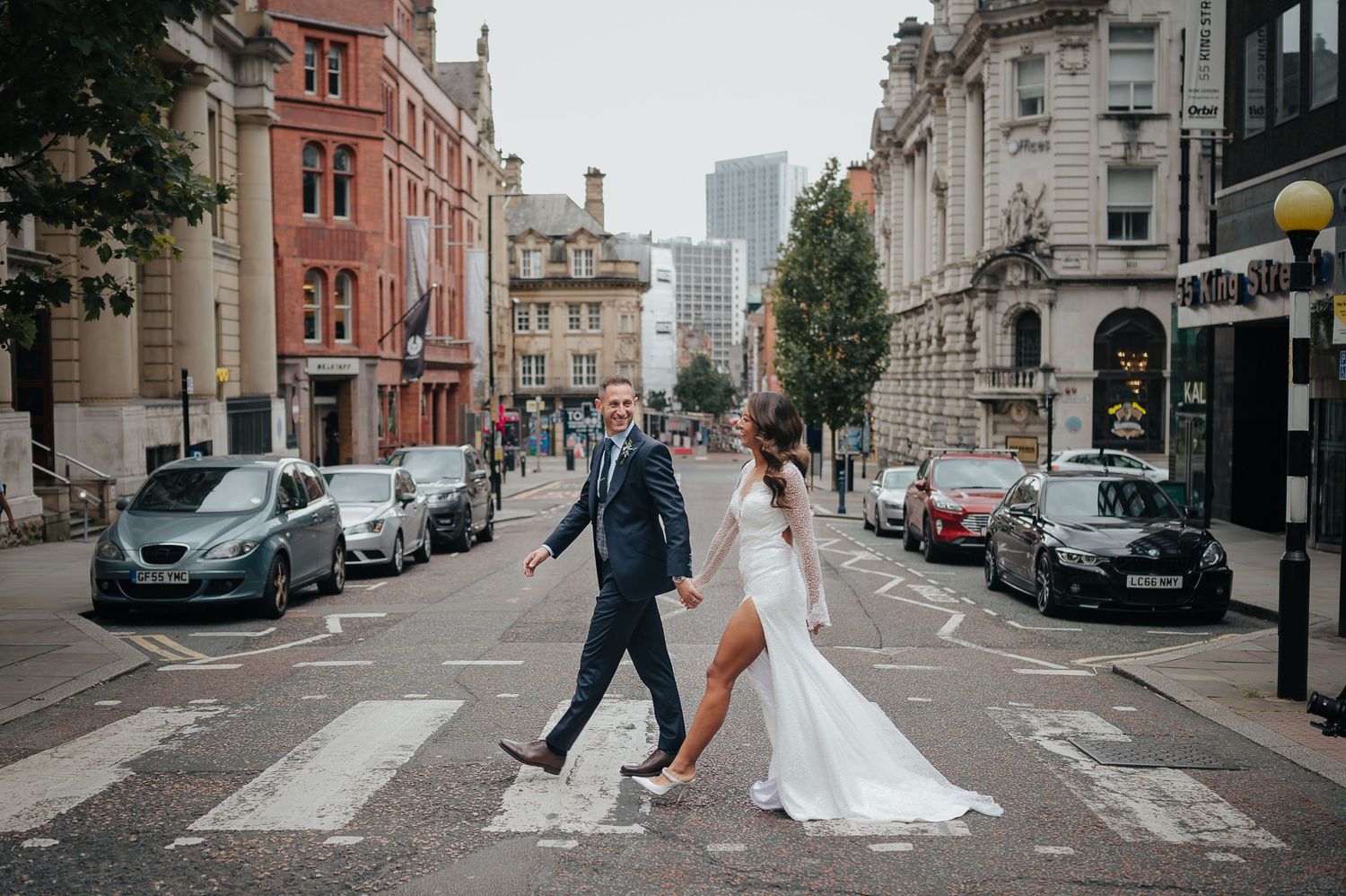 Newlyweds recreate iconic Beatles Abbey Road crossing pose in urban setting with vintage architecture.