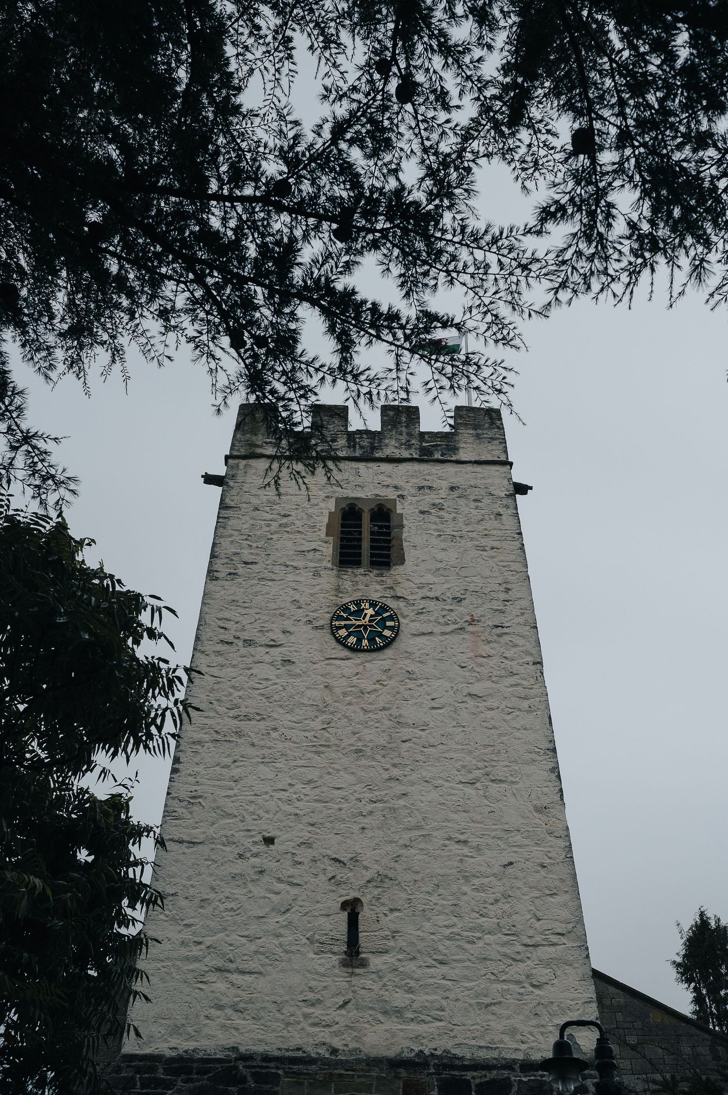 A white stone church tower with clock face framed by overhanging tree branches.