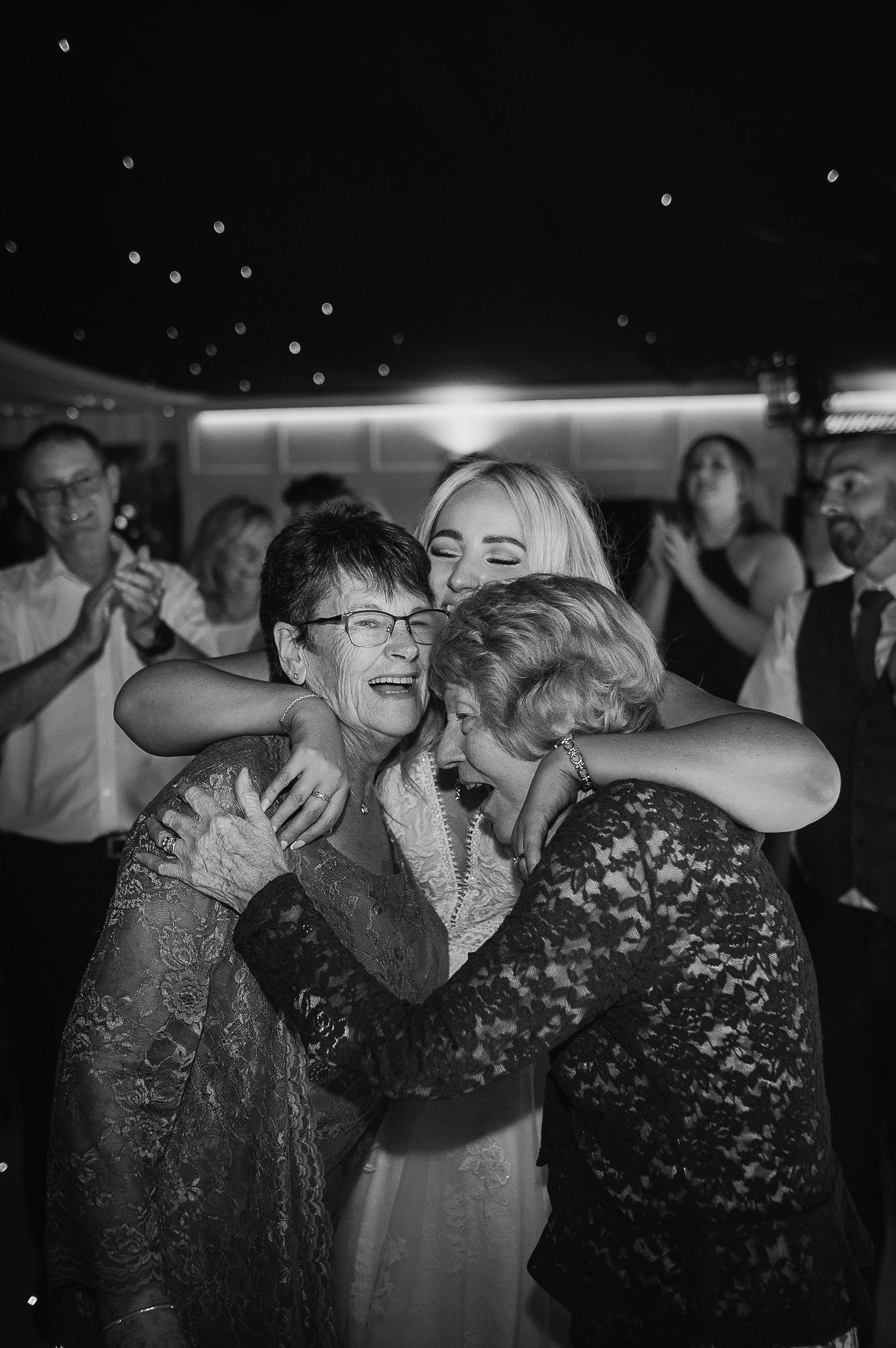 Black and white photograph capturing emotional group hug during wedding reception celebrations.