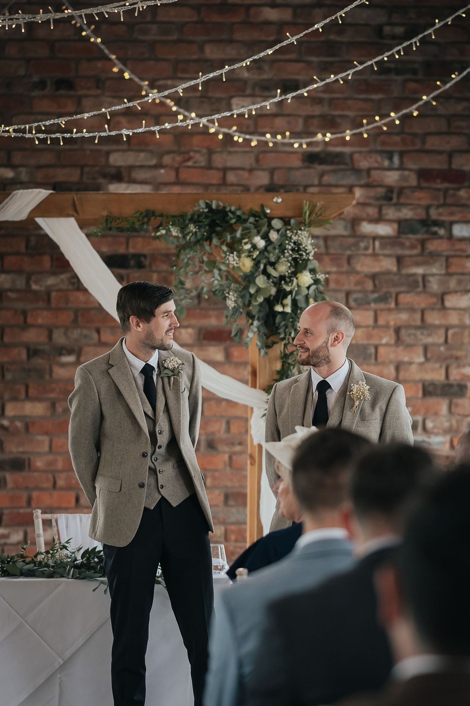 Wedding guests in suits gather under string lights and floral decorations against a brick wall.