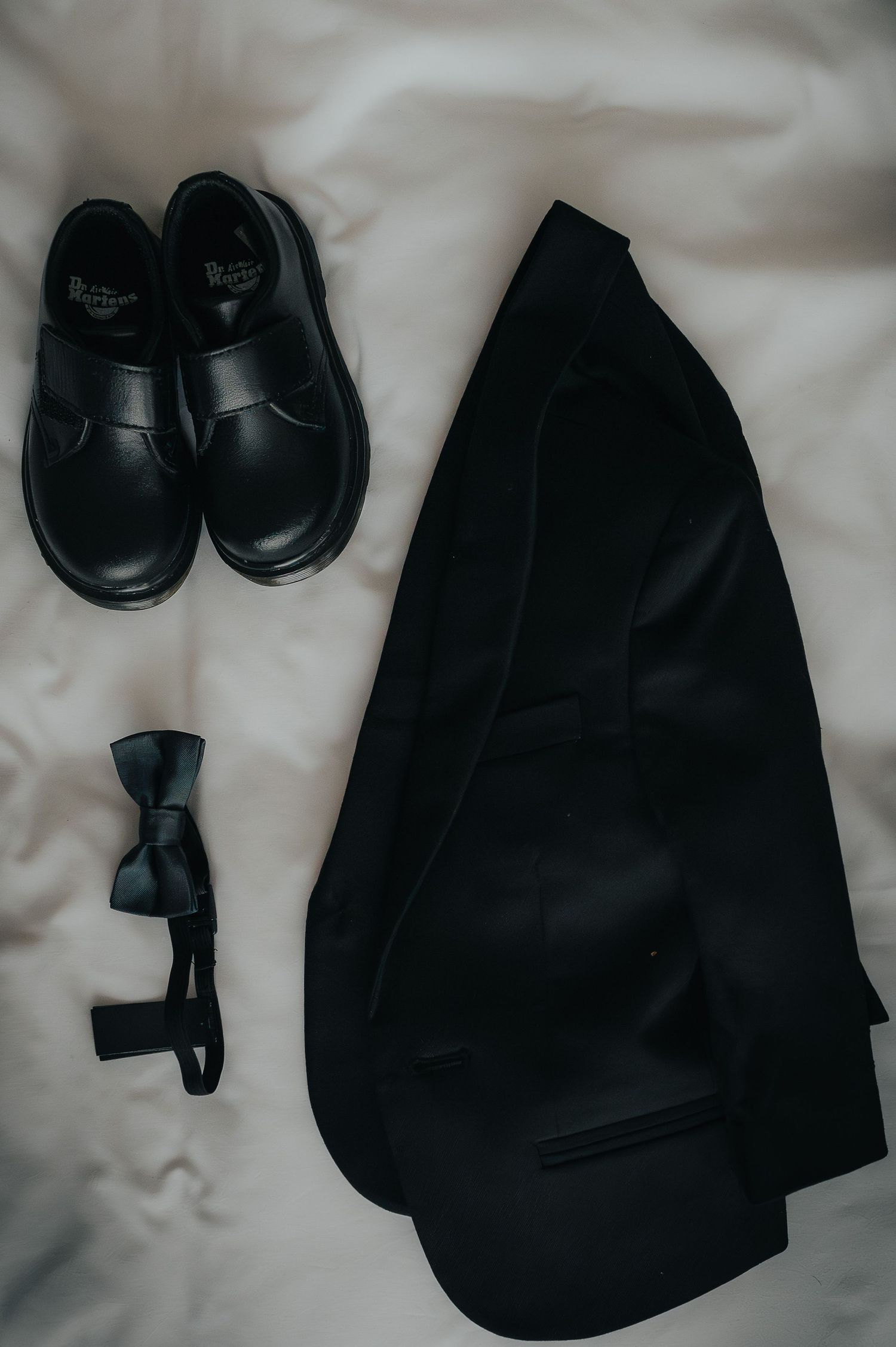 A black school uniform set laid out on white fabric, featuring dress shoes, a bow tie, and a blazer jacket.