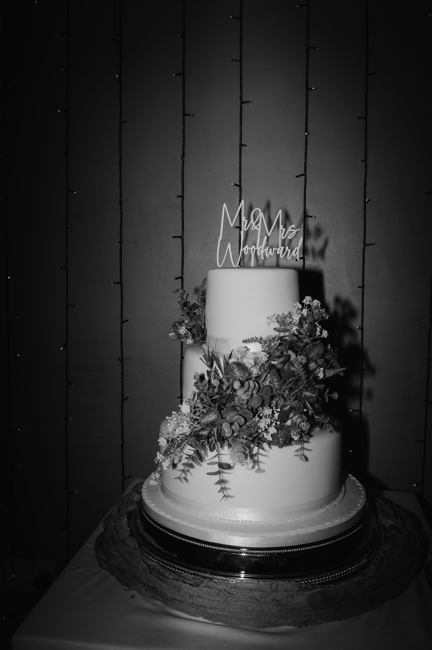 Elegant white wedding cake decorated with greenery and a neon sign topper on a dark background.
