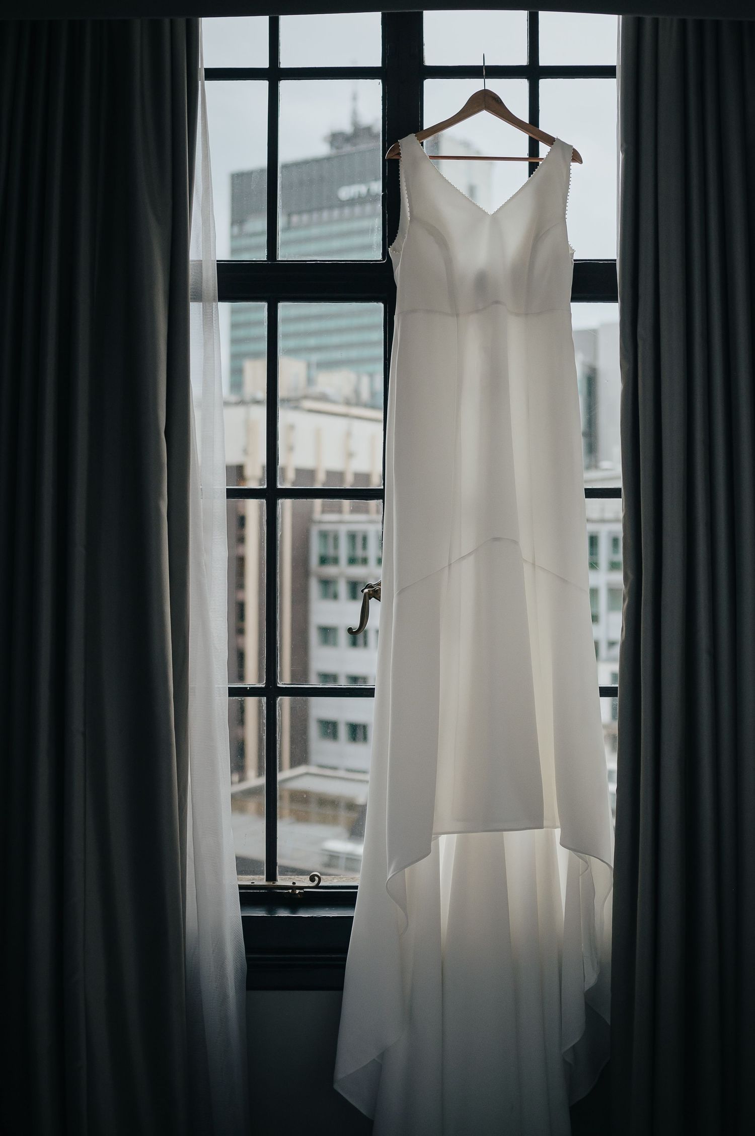 A white wedding dress hangs in silhouette against a window with dark curtains.