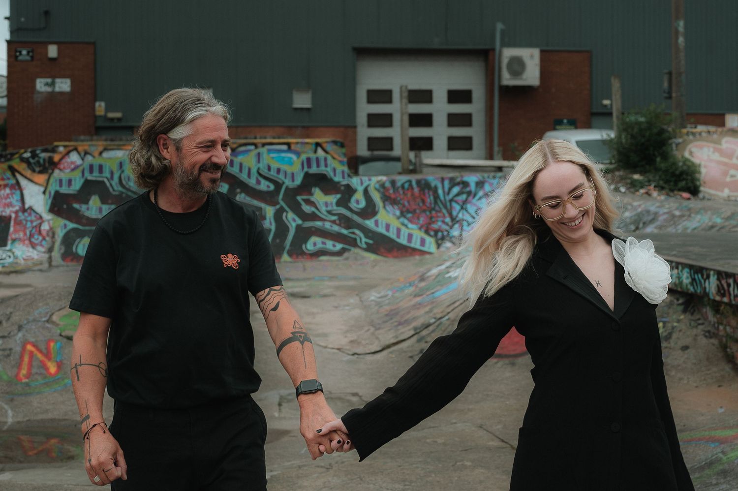 People in black clothing dance and move joyfully in an urban setting with graffiti-covered walls.
