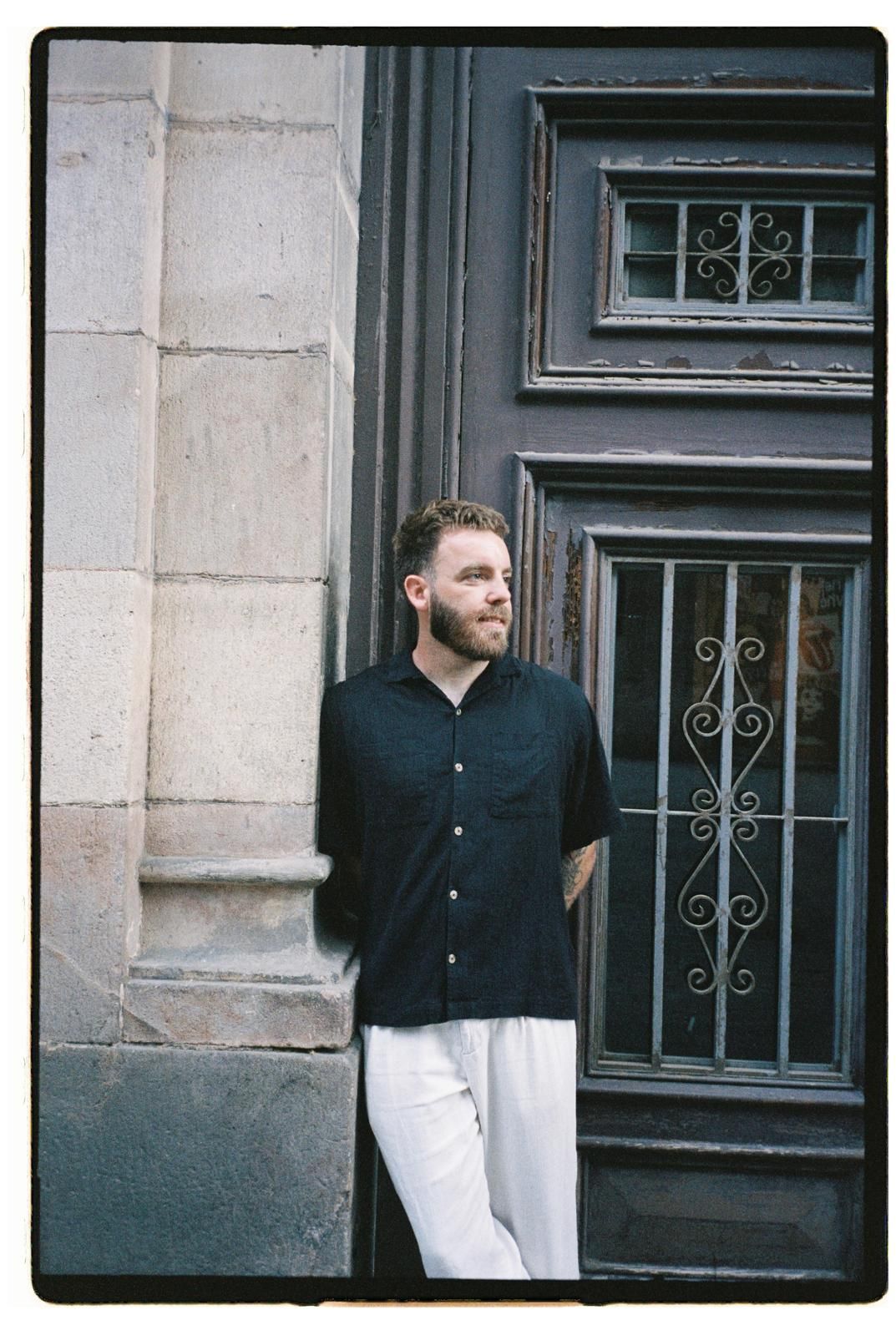 Man in black shirt and white pants leaning against a stone wall beside a vintage dark wooden door.