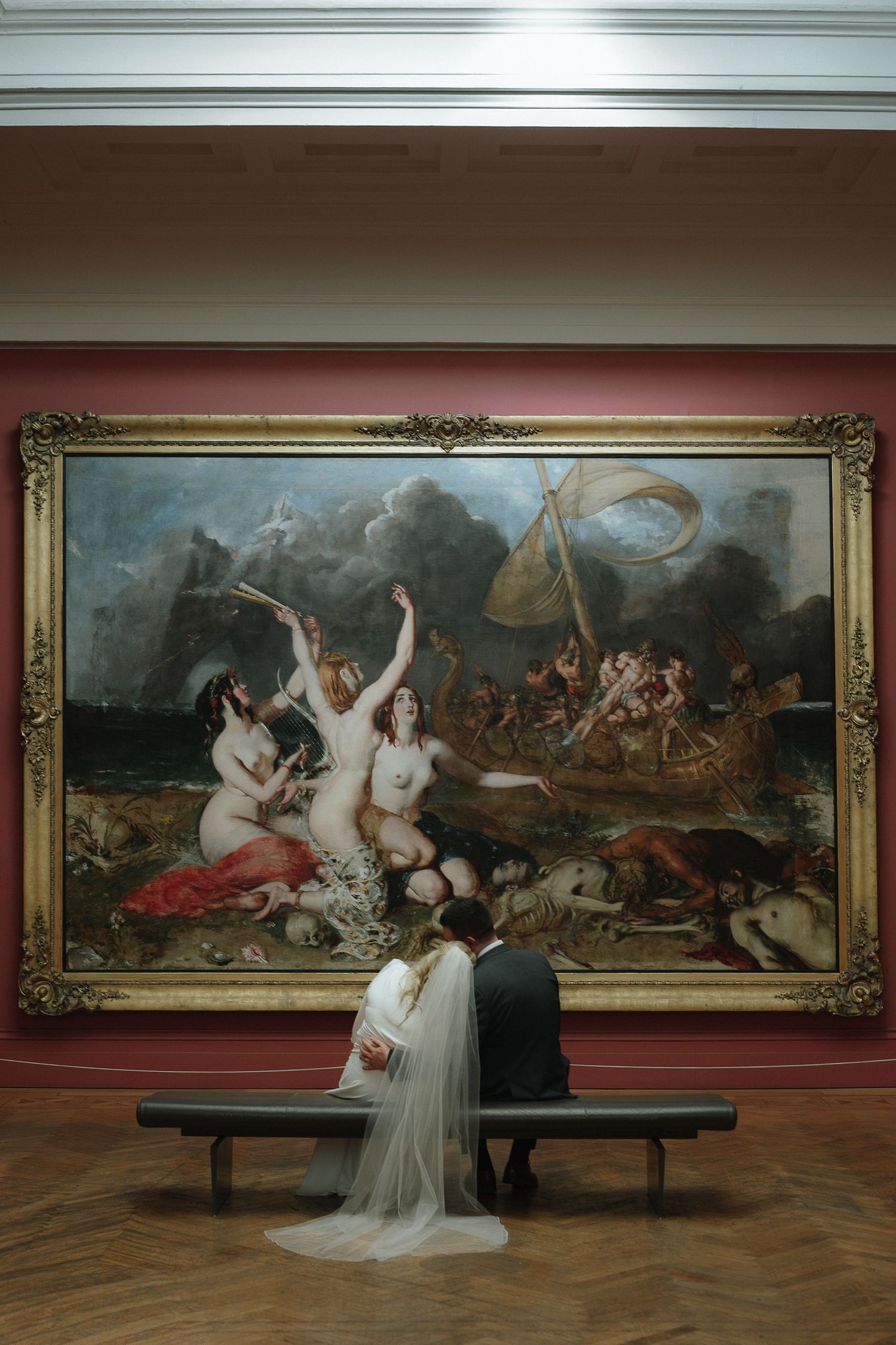 Bride and groom sitting on bench before large classical painting in art museum wedding photo. Manchester Wedding Photographer.