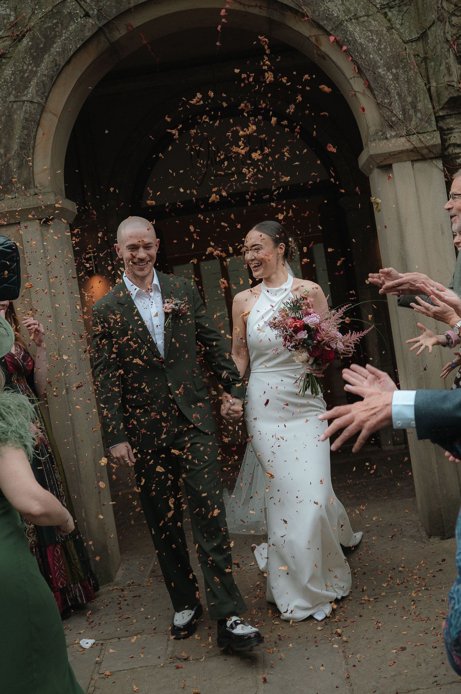 London Wedding Photographer. Newlywed couple walking through confetti shower outside stone church, bride in white gown holding bouquet,