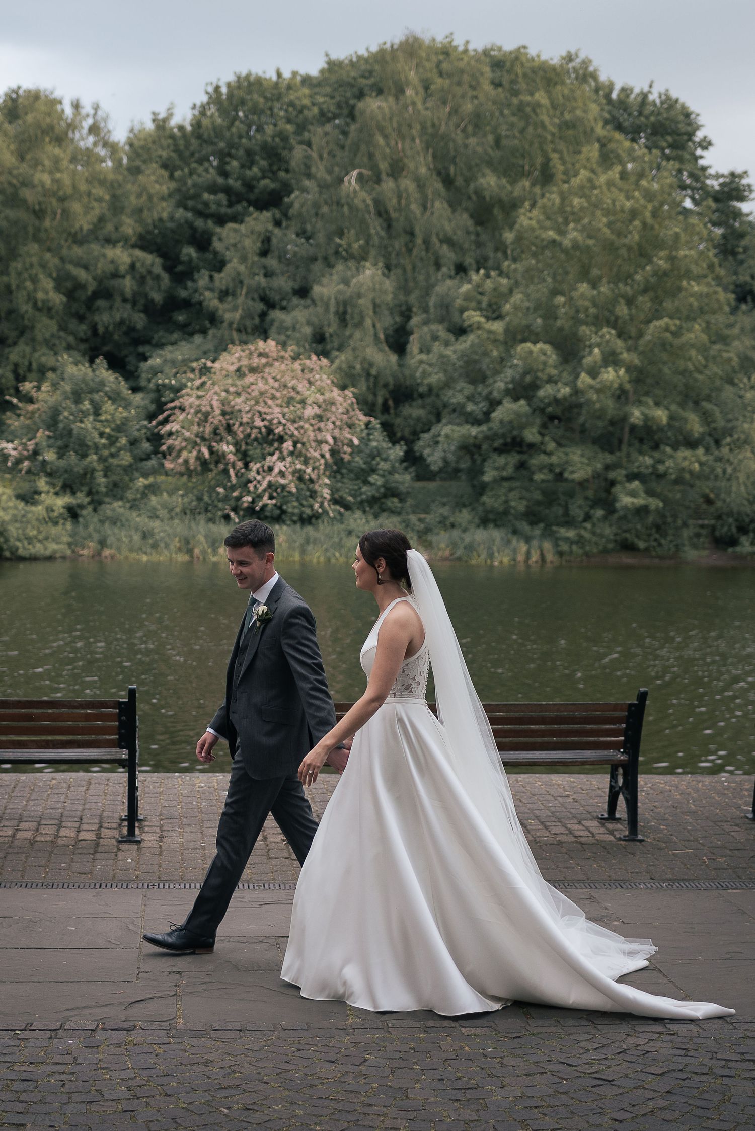 Couple strolling beside a serene lake with lush greenery in the background.