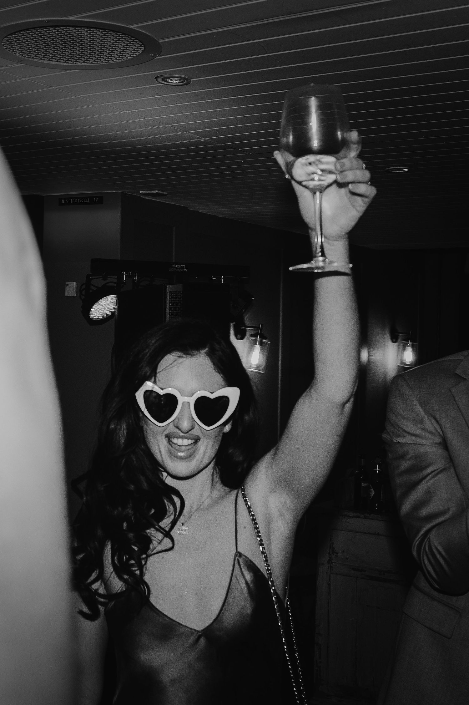 Black and white photo of party guest wearing heart sunglasses while raising glass in celebration.
