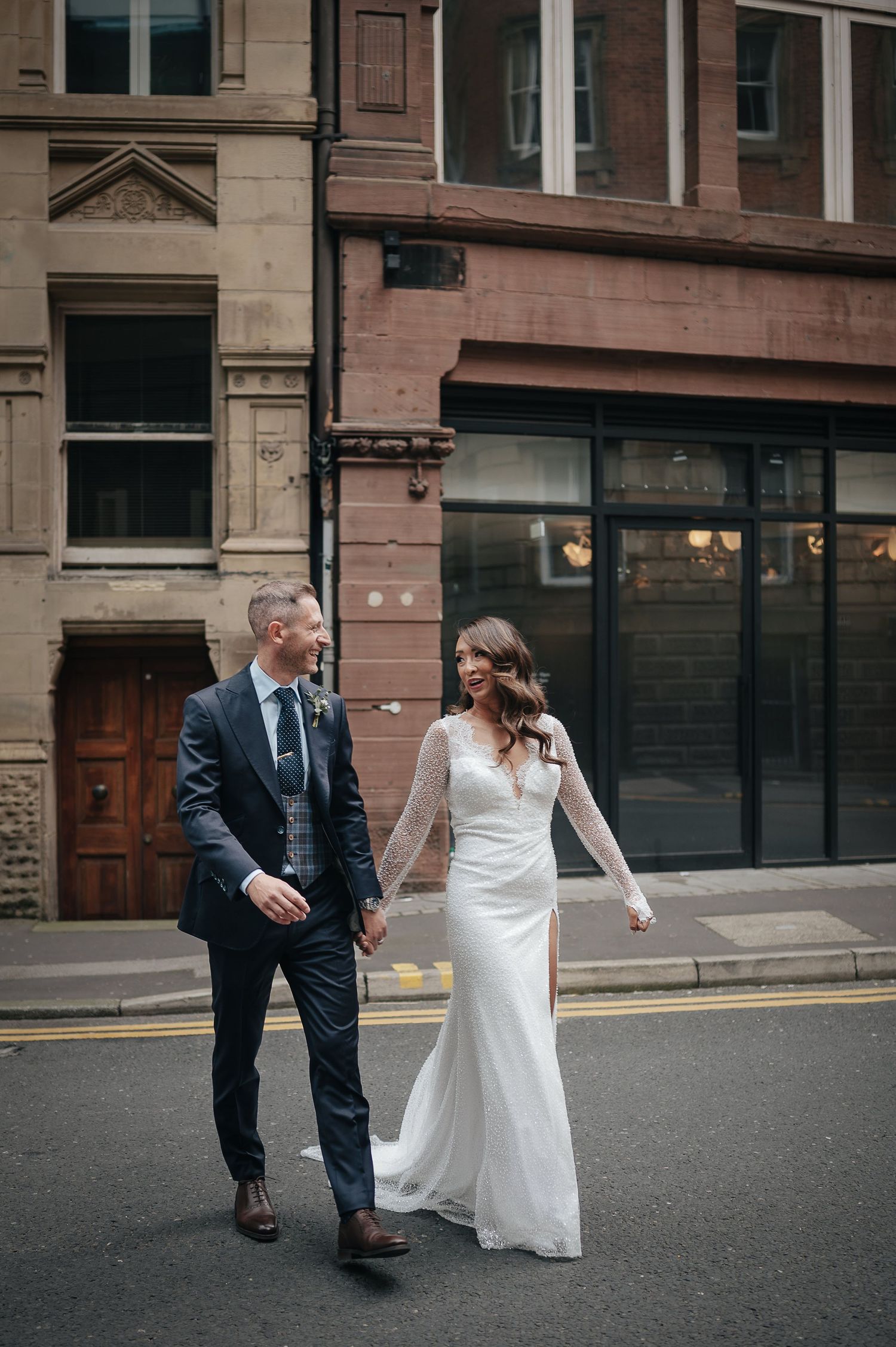 Elegant couple walking down urban street in wedding attire against brick buildings.