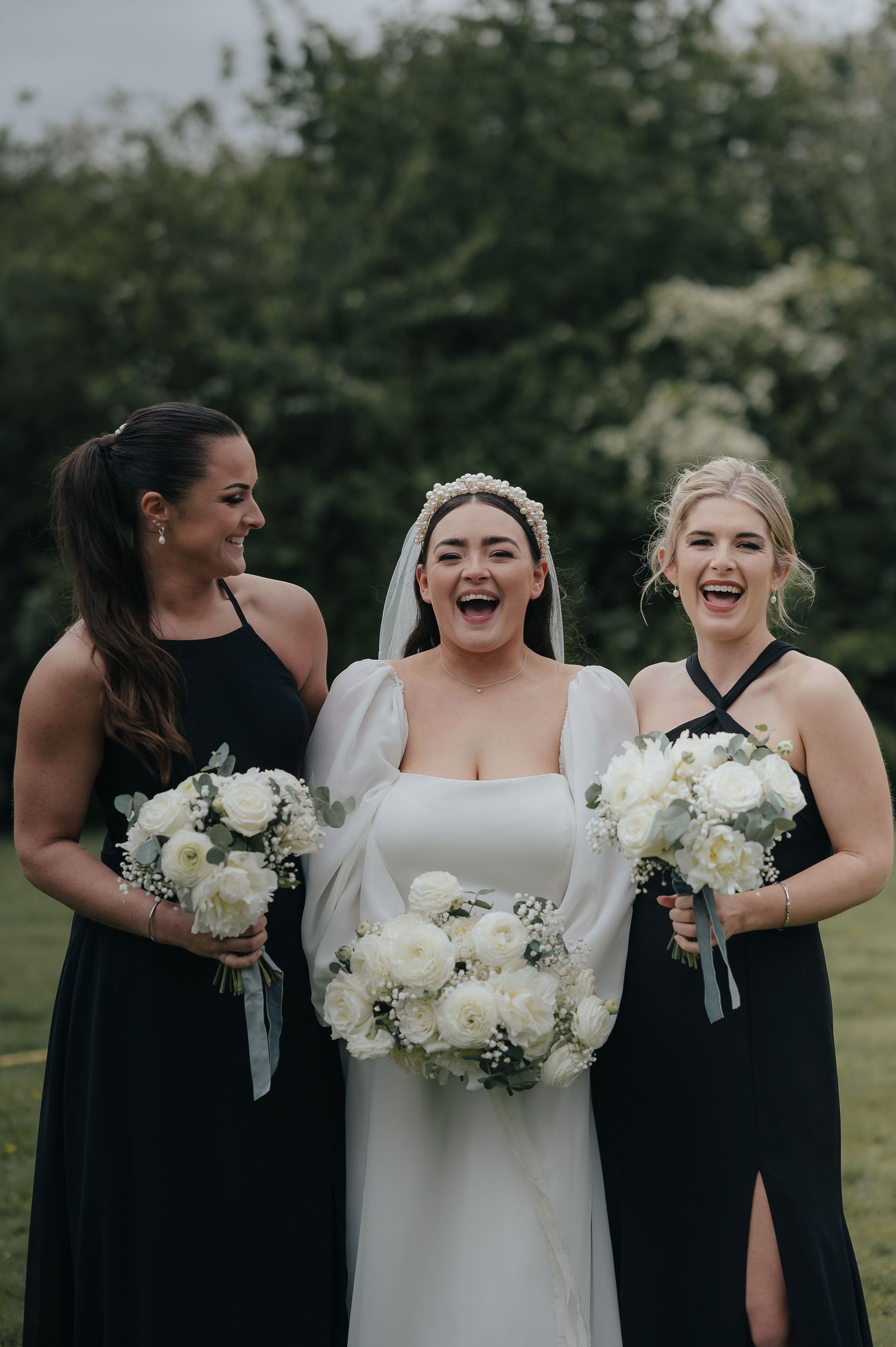 Three women in formal attire hold white bouquets while laughing together outdoors.