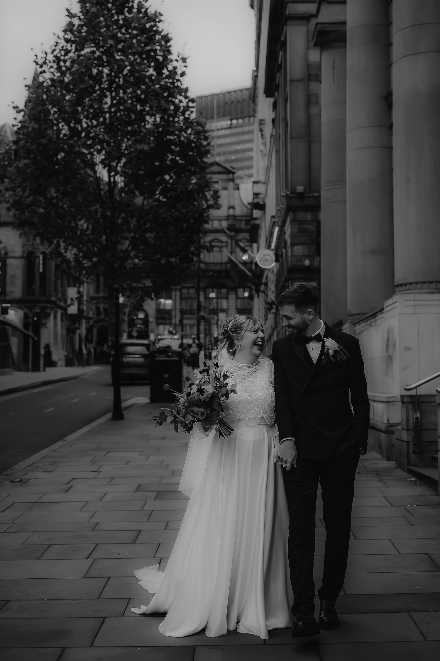 A newly married couple stands on a city sidewalk with classic architecture and trees in black and white.
