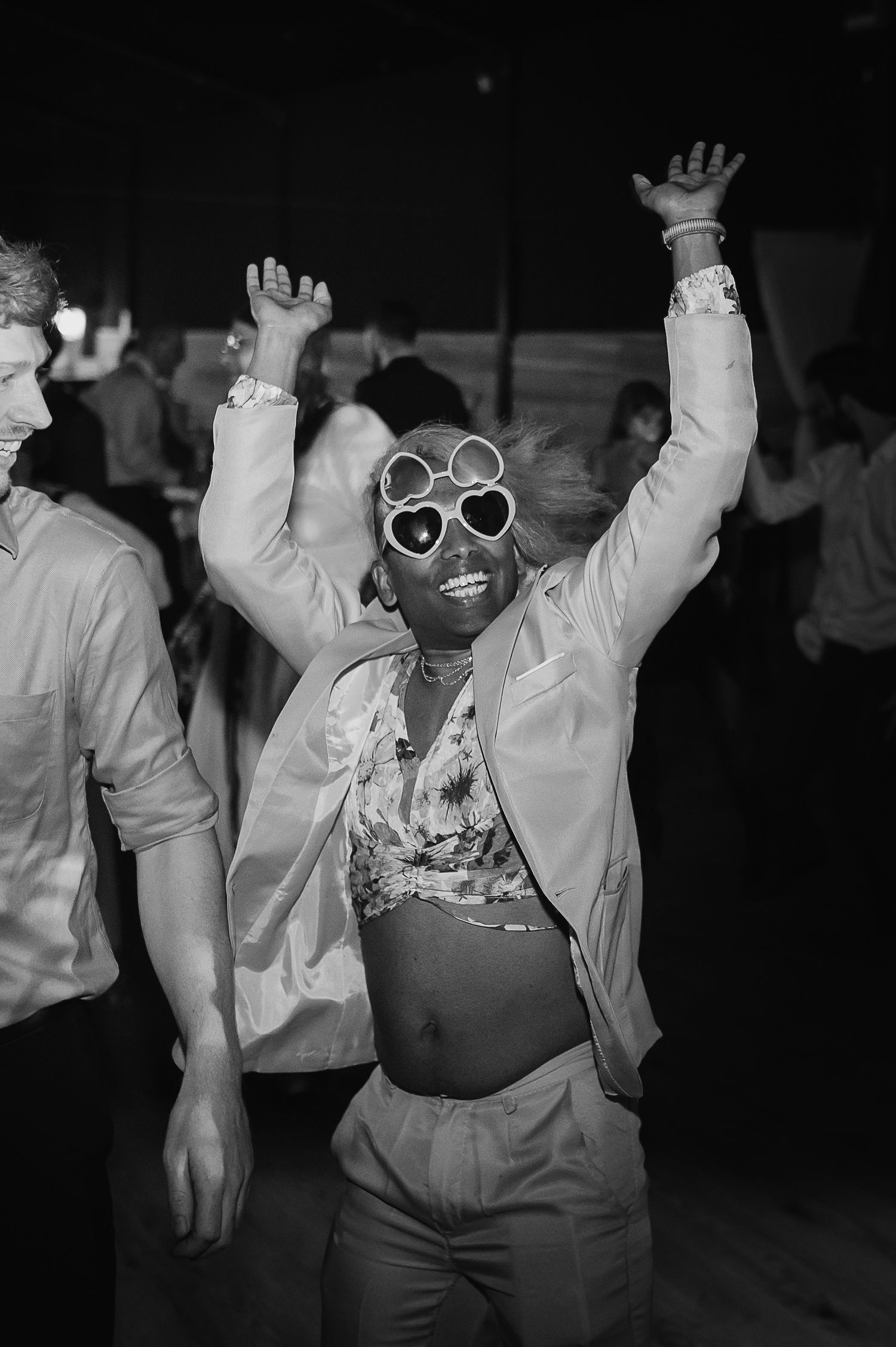 Dancer in heart-shaped sunglasses and white jacket celebrating on dark dance floor.