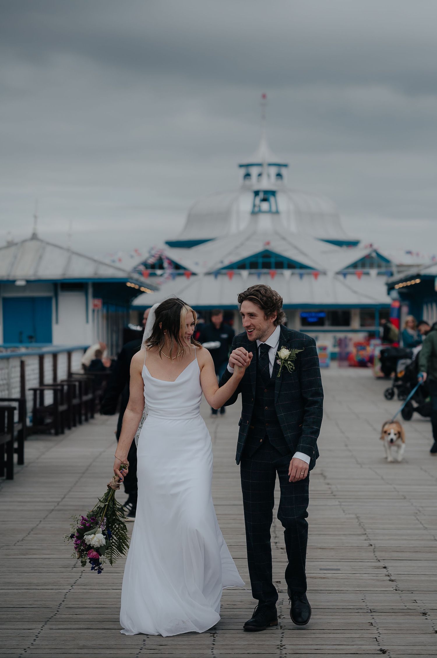 A couple strolling down a pier with Victorian architecture and a small dog in the background.