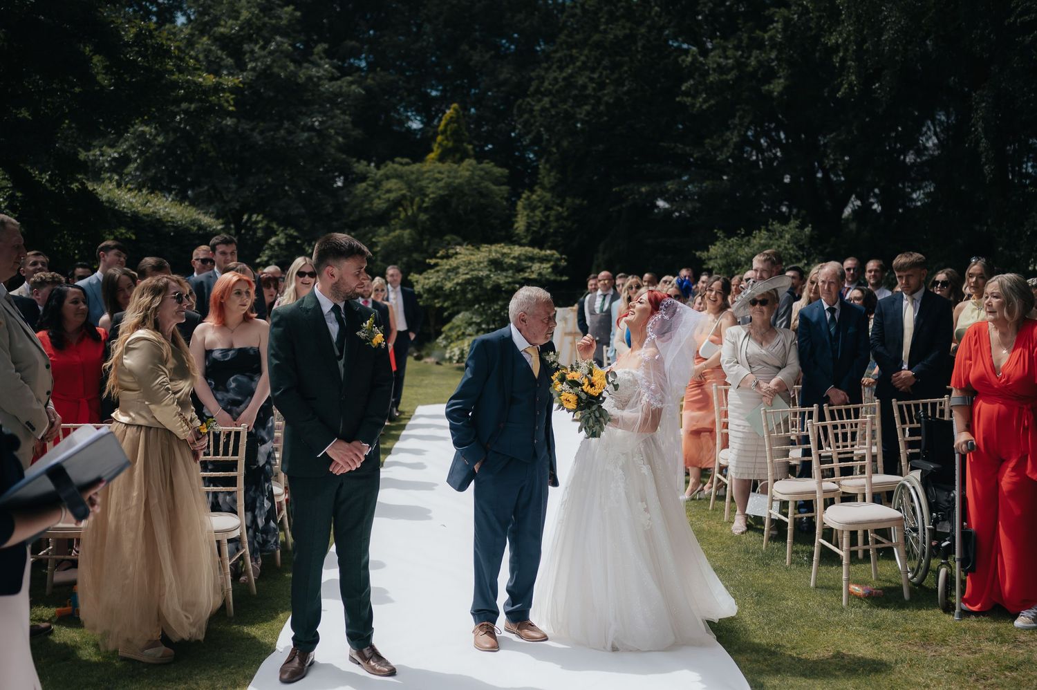 Outdoor wedding ceremony with guests seated on white chairs along a white aisle runner in a garden setting.