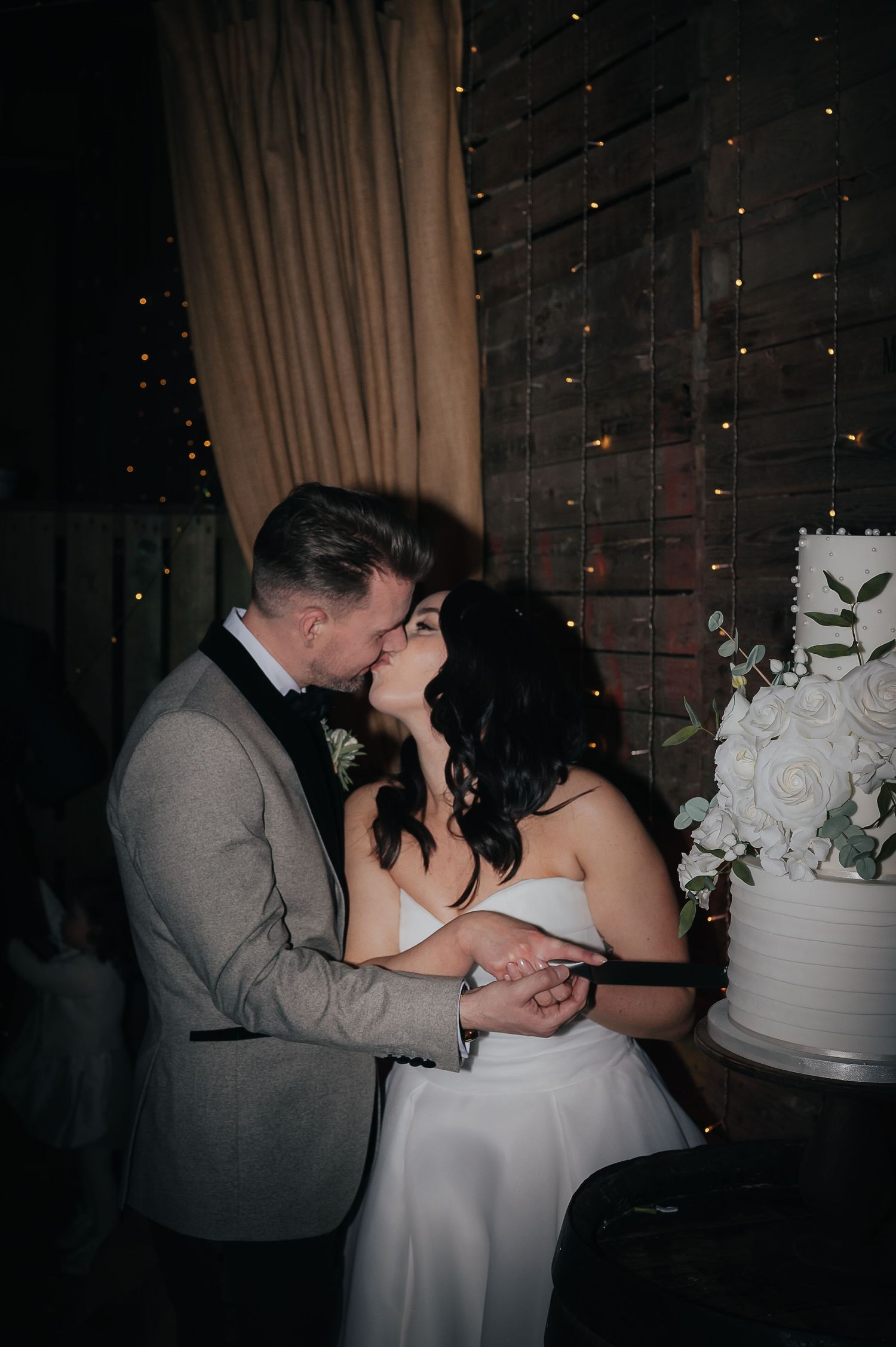 An intimate moment between a couple next to their white wedding cake under string lights.