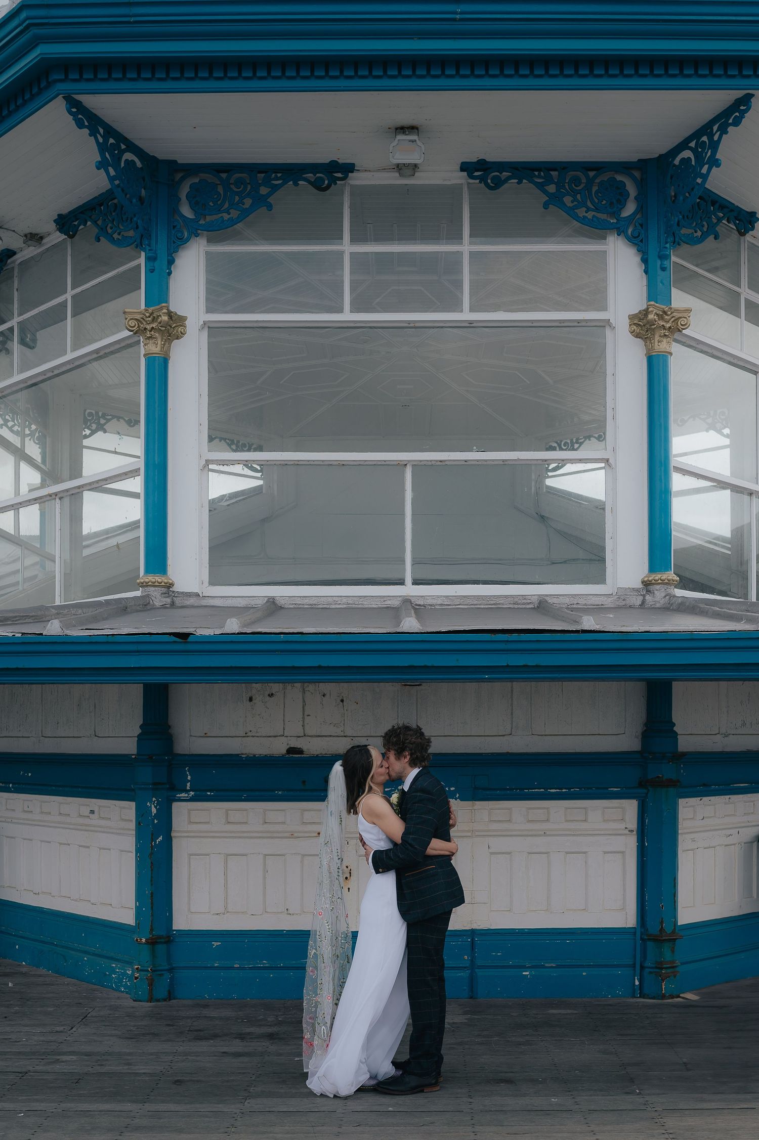 A romantic wedding moment under an ornate blue and white Victorian-style bandstand structure.