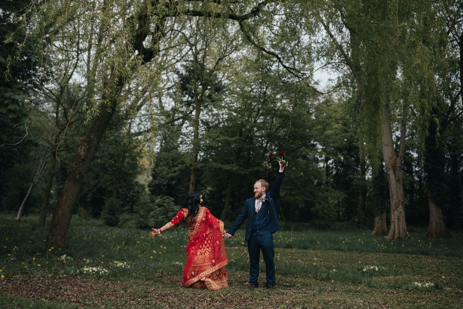 A couple dances in a wooded area, with one wearing a vibrant red dress against green foliage.
