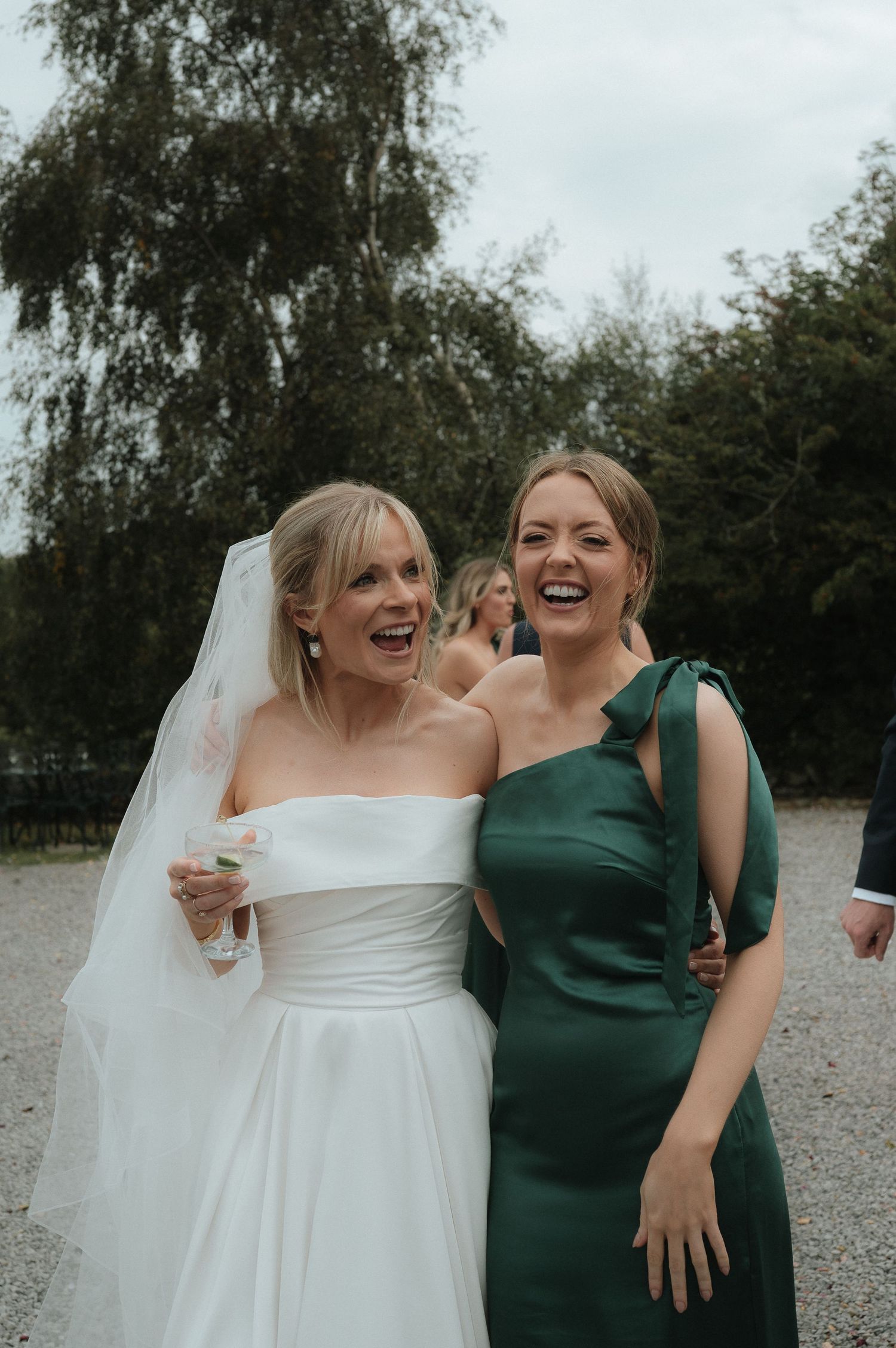 A bride in a white dress and her friend in emerald green share a candid laughing moment at a wedding.
