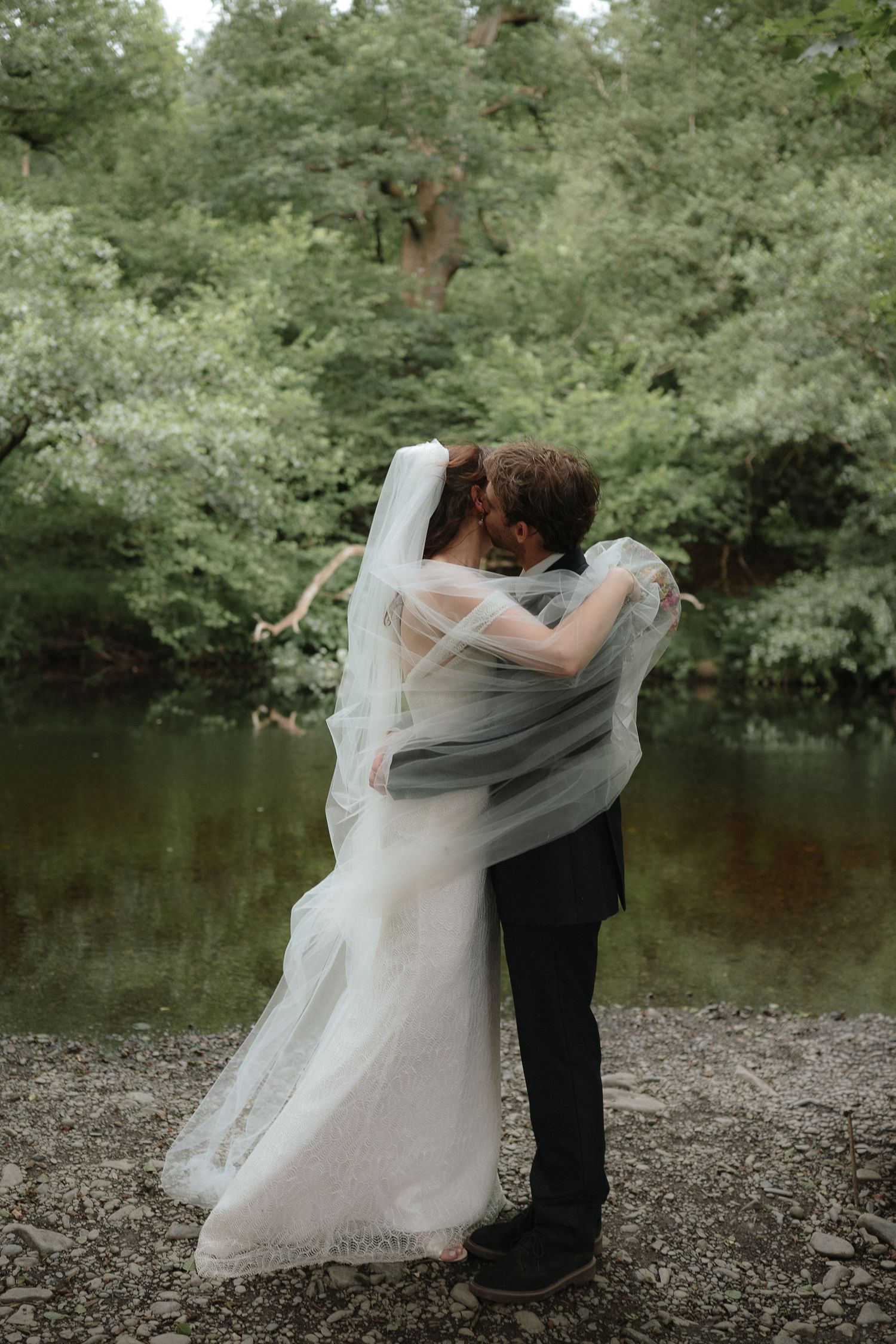 Bride and groom sharing a romantic kiss by a serene lake, surrounded by lush green trees, with a flowing white veil.