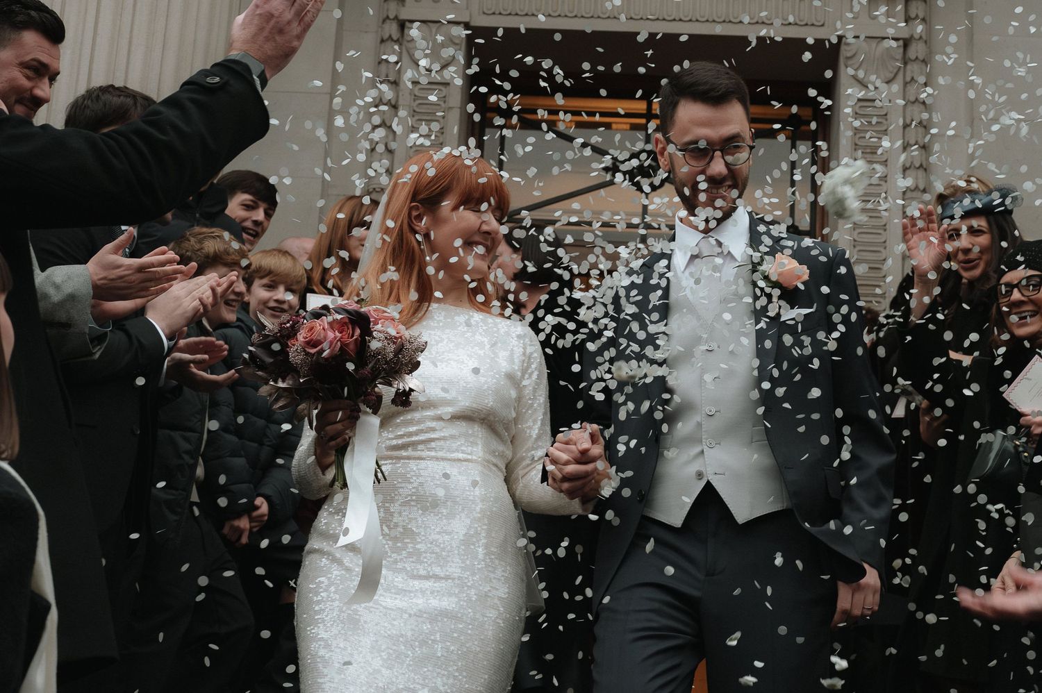 Bride and groom walk through confetti shower surrounded by cheering guests outside wedding venue. London Wedding Photographer.