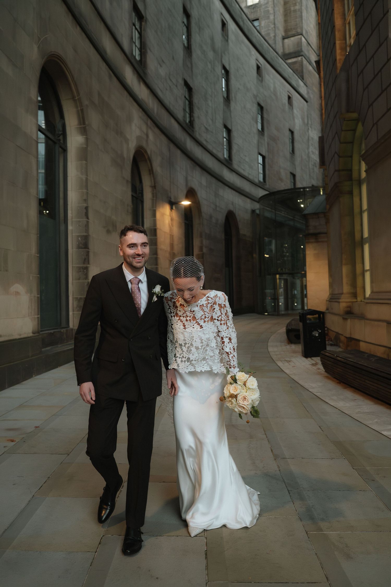 A smiling groom in a dark suit walks with his bride in a lace-topped wedding dress holding a bouque. Manchester Wedding Photographer.