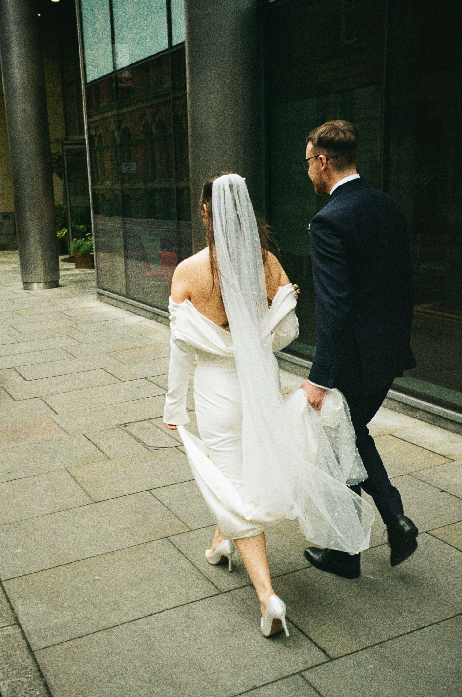 Bride in white gown and veil walks with groom in navy suit along urban pavement outside modern building.