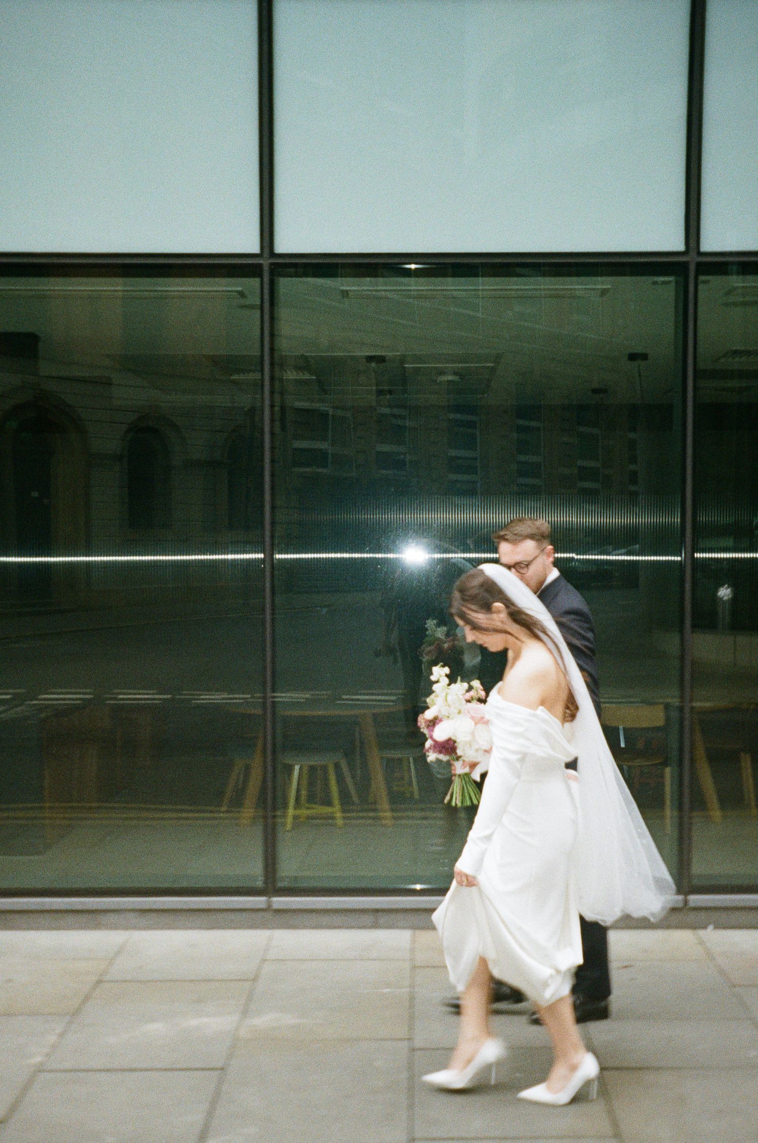 Bride and groom walking past modern glass building, bride in off-shoulder white gown holding floral bouquet.
