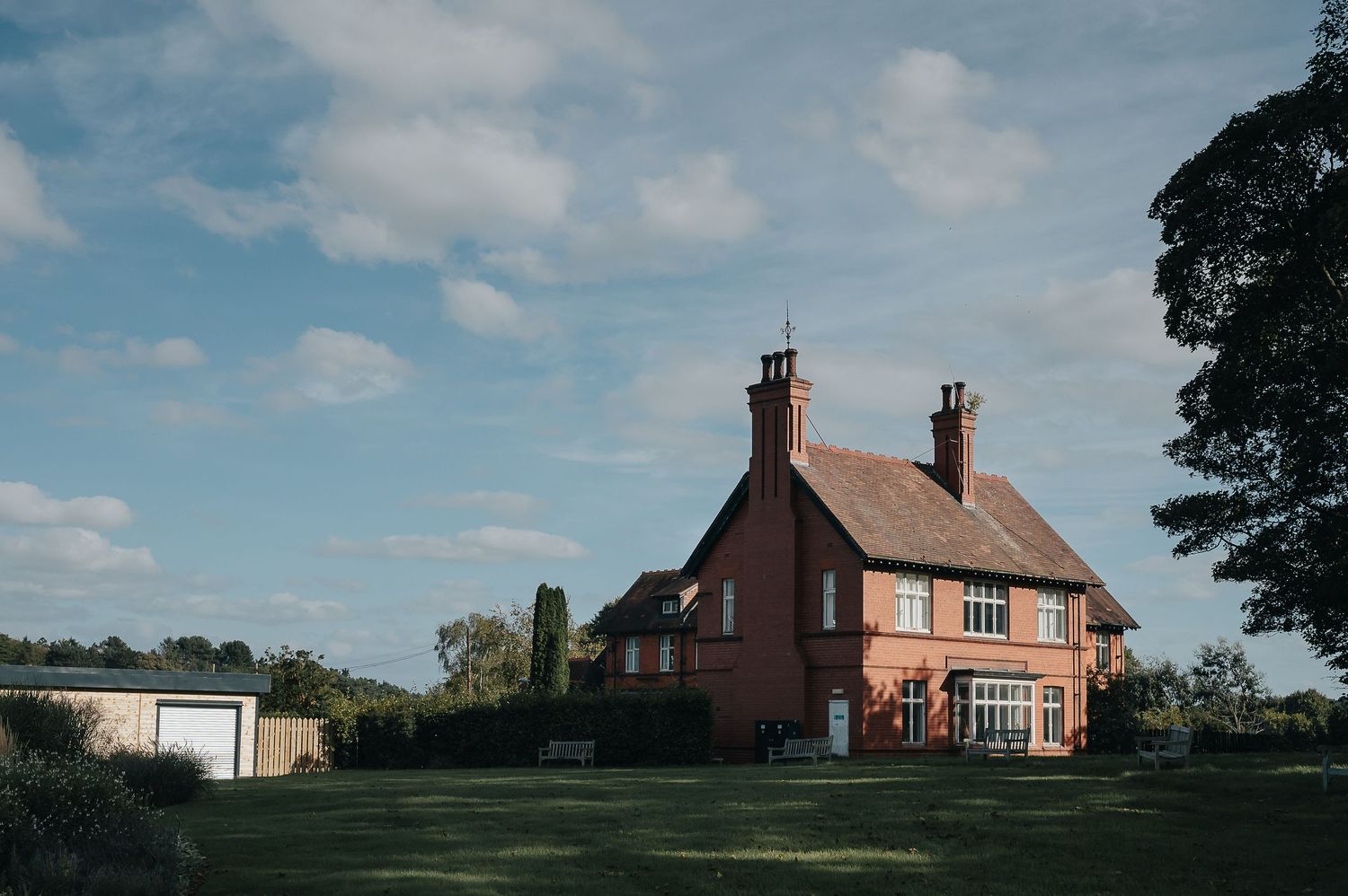 A red brick country house stands on a green lawn under a bright blue sky with white clouds.