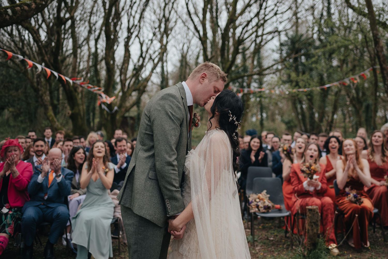 Outdoor woodland wedding ceremony with guests watching couple share their first kiss.