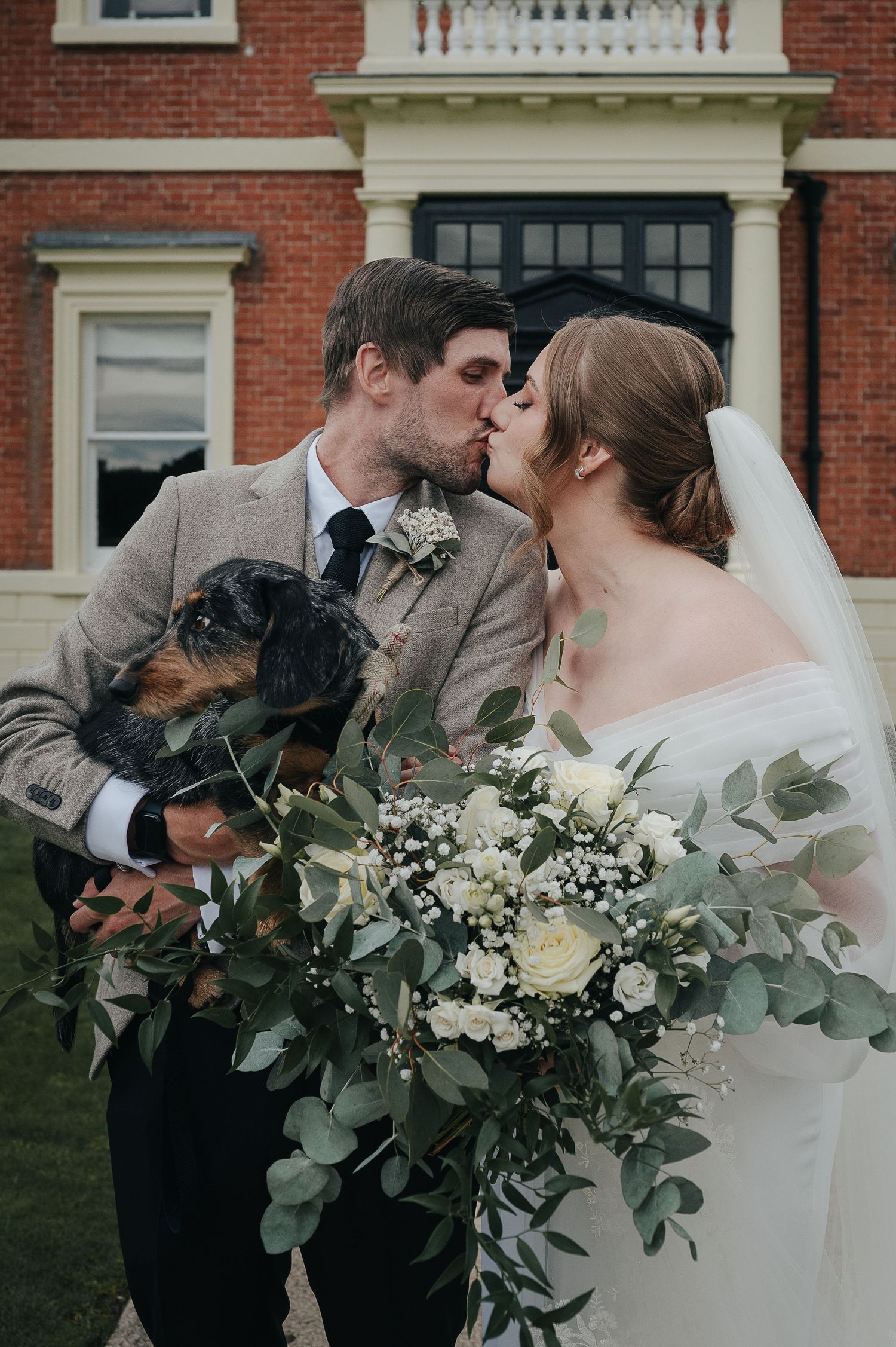 A romantic wedding portrait with a couple and their small dog holding a lush greenery bouquet.