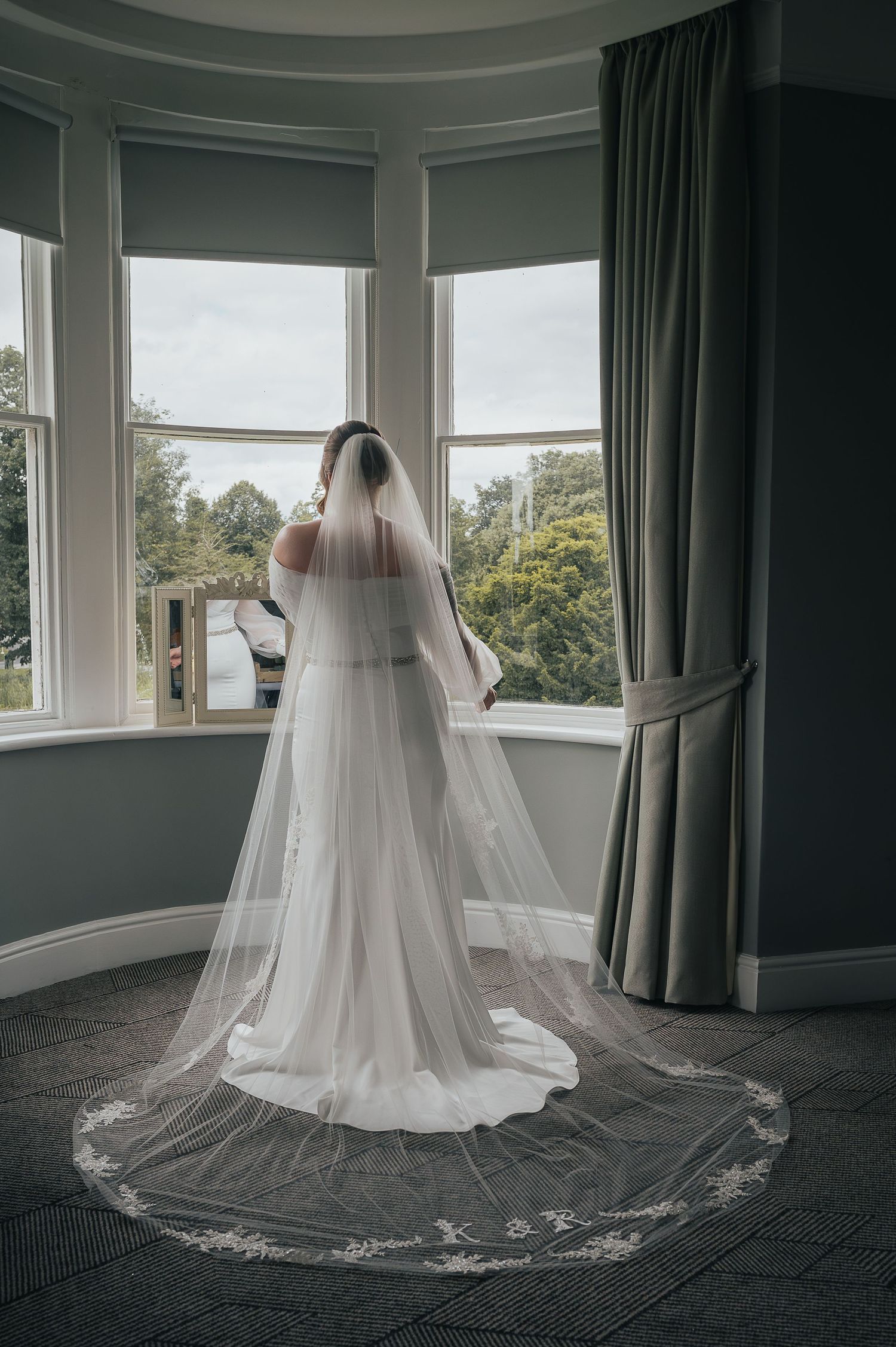 Elegant wedding dress and cathedral veil photographed from behind in a bay window setting.